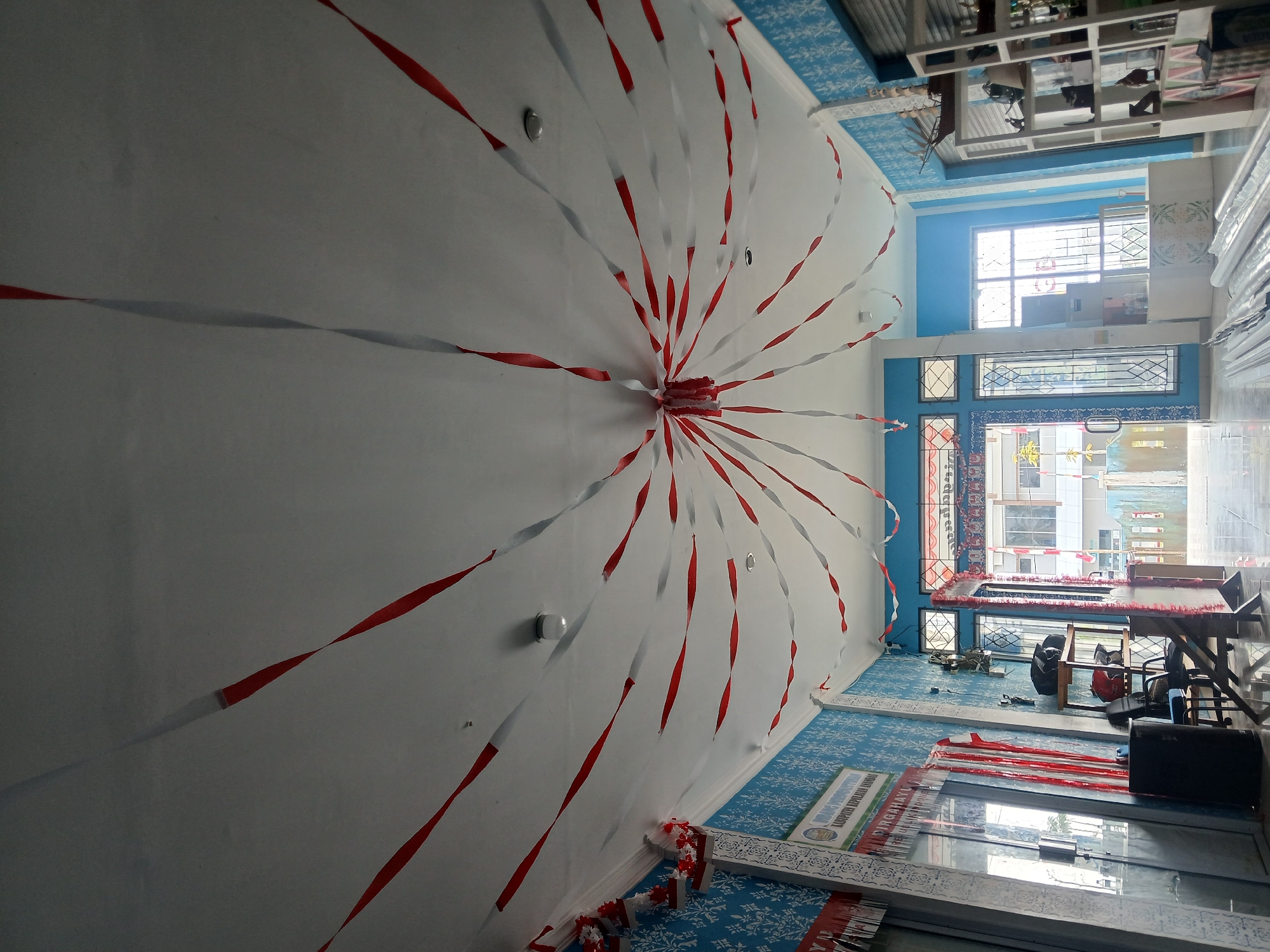 The image depicts the interior of a building, likely a public space or office, decorated with red and white streamers. The streamers are suspended from the ceiling, creating a radial pattern emanating from a central point. The walls are painted blue with a white patterned wallpaper. In the foreground, a tiled floor is visible, with some rolled-up material placed on it. To the left, a glass door and a black trash can are present. Further back, there is a doorway leading to an area with more windows and what appears to be a stage or platform. Shelving units with various objects are visible on the right side of the image. The overall impression is one of festive decoration, possibly for a national holiday given the red and white color scheme, which are the colors of the Indonesian flag. The building is located in Tarempa Timur, Indonesia, as indicated by an inscription on a sign to the left of the image which reads "PEMERINTAH KABUPATEN KEPULAUAN ANAMAS DIRGAHAYU".