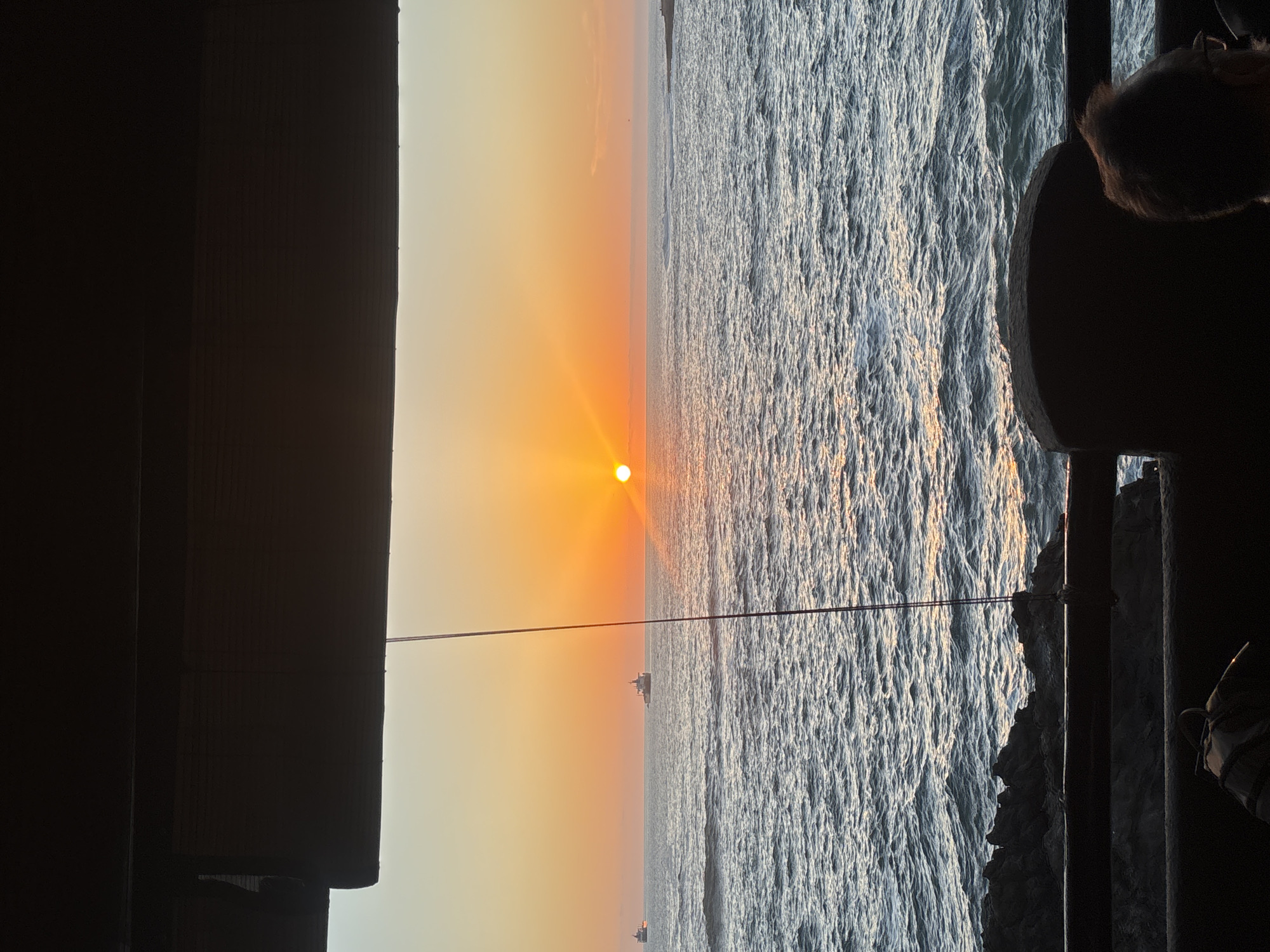 A landscape photograph shows a sunset over the ocean in Arica, Chile. The sun is low on the horizon, casting an orange glow over the sky. The ocean is calm and reflects the sunlight. The photograph is taken from an elevated location on a structure, with portions of the structure visible on both the left and right of the photo. There are small boats visible on the horizon line.