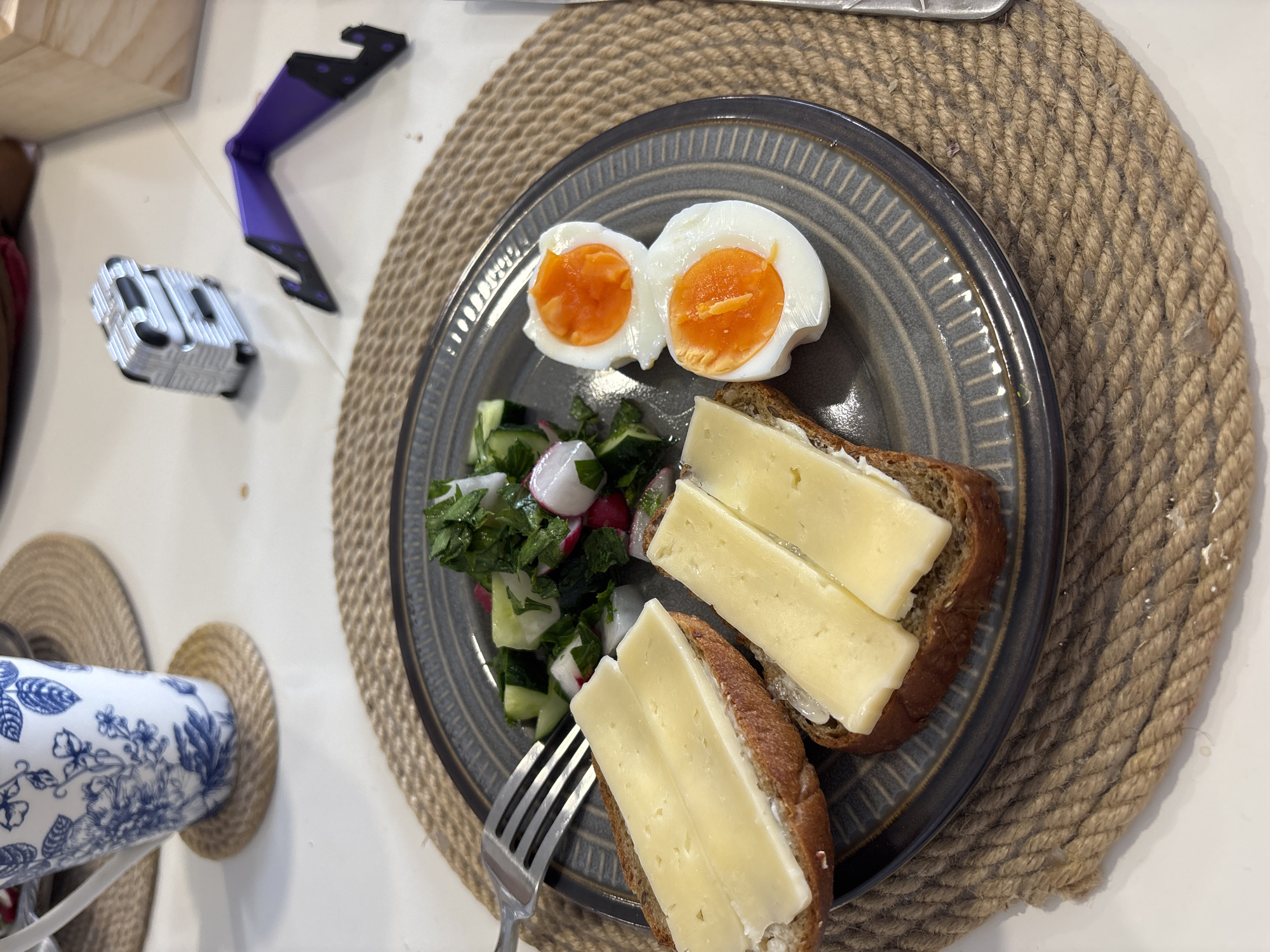 A plate of breakfast food sits on a woven placemat. The plate contains two pieces of rye bread topped with cheese, a side salad of cucumber, radish, and greens, and two halves of a soft-boiled egg with vibrant orange yolks. A fork rests on the edge of the plate. In the background, a blue and white patterned mug sits on another woven placemat. A small, silver suitcase-shaped object and a purple phone stand are also visible on the table. The overall setting appears to be a domestic dining area. There is no information in the image to suggest the location is Krasnoyarsk, Russia.