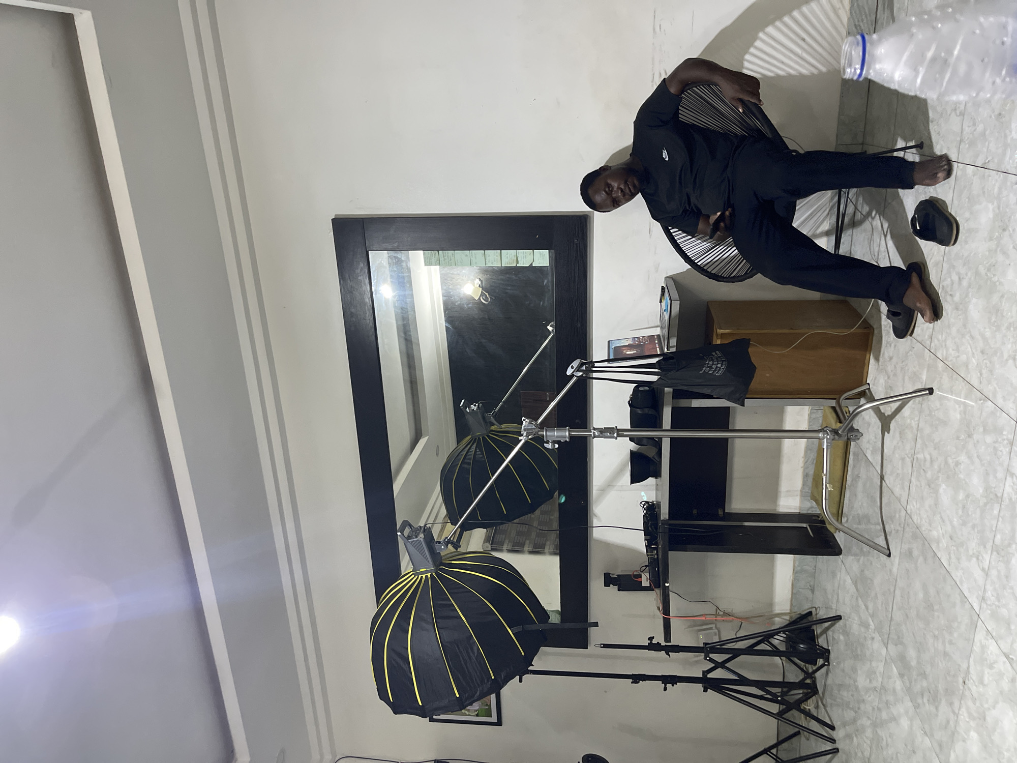The image displays an indoor scene, likely a photography studio or a dedicated space for photoshoots, located in Abuja, Nigeria. A man is seated on a black, slatted chair, facing the viewer with a neutral expression. He is wearing a black short-sleeved shirt featuring a white Nike logo on the left chest, and black trousers. His bare feet are visible, with a pair of dark sandals resting on the light-colored tiled floor beside them. A clear plastic water bottle is also on the floor nearby.
The room features white walls and a reflective, light-toned tiled floor. A significant portion of the image is occupied by a large, dark-framed mirror positioned against the wall, which reflects other elements of the room, including photography equipment. In the foreground, a black parabolic softbox with yellow internal stripes is set up on a light stand. Another similar softbox and a C-stand are visible in the mirror's reflection, suggesting a two-light setup. A powerful artificial light source, located out of frame to the left, casts a bright flare into the lens, illuminating the scene.
Behind the seated man, a wooden cabinet or desk holds various electronics, possibly a laptop or tablet, and tangled cables. A black bag is on the floor next to this area. The overall arrangement points to a professional or semi-professional photography session taking place, utilizing specialized lighting equipment.
