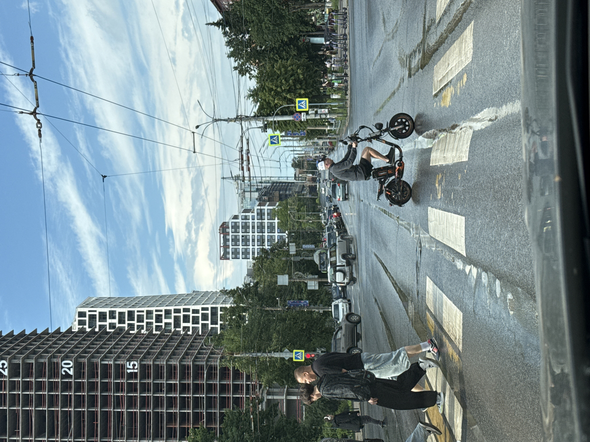 The image depicts a street scene in Izhevsk, Russia, likely near a construction site given the unfinished high-rise buildings in the background. The focus is on a crosswalk where several individuals are crossing. A man on an electric scooter is in the process of crossing the street. In the foreground, two pedestrians are walking across the crosswalk, one in black pants and a patterned top, the other in a black t-shirt and light blue shorts. Behind them, another person in black pants and a black jacket with headphones is also visible on the crosswalk. The street is wet, indicating recent rain. Several cars are visible on the road, and overhead power lines suggest a tram or trolleybus system is in operation. The sky is partly cloudy with a blue hue. Traffic signals and pedestrian crossing signs are present. The overall impression is a typical urban street scene with active pedestrian and vehicle traffic.
