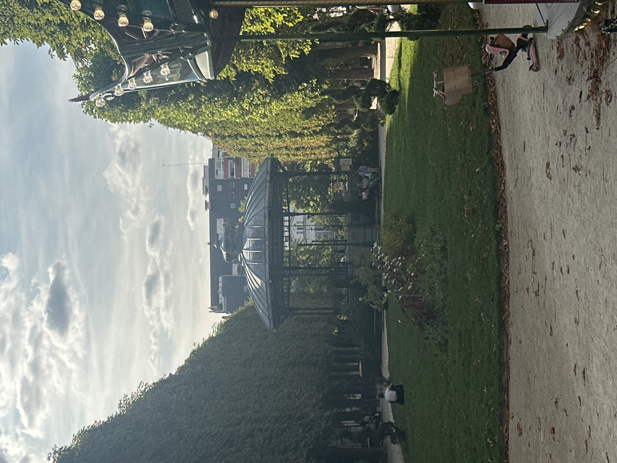 The image captures a park scene in Paris, France, likely during the daytime. In the center, a black, ornate gazebo with a domed roof stands on a grassy area. It is surrounded by mature trees with dense foliage, which form archways or corridors on either side of the central lawn. Several people are seated on the grass or on benches near the gazebo. In the foreground, a gravel path runs horizontally. To the right, a portion of a carousel is visible, with its decorative lights and structure partially obscuring the trees. The sky above is partly cloudy. The overall atmosphere suggests a peaceful, public space within the city.