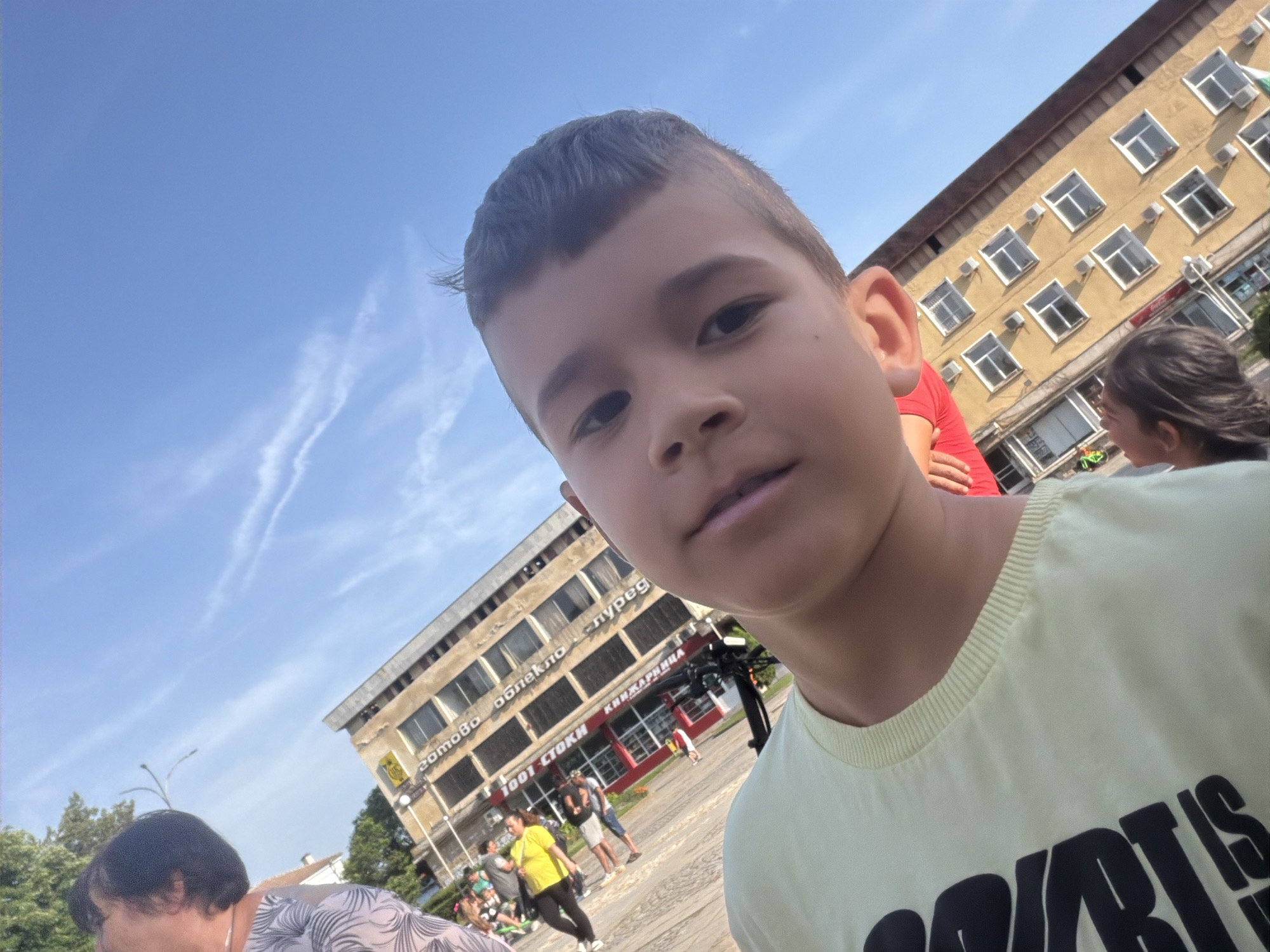 A young boy is taking a selfie outdoors on a sunny day in the city of Unknown. The sky is blue with faint contrails. In the background, two buildings are visible. One is a light yellow, multi-story building with numerous windows, and a sign in a non-English language. The other building is a similar design. People are walking around in the plaza between the buildings. To the left of the frame is an older woman with short dark hair and a patterned shirt. To the right of the frame is a girl with dark hair and a red-shirted individual. The boy is wearing a light yellow t-shirt with large black letters.