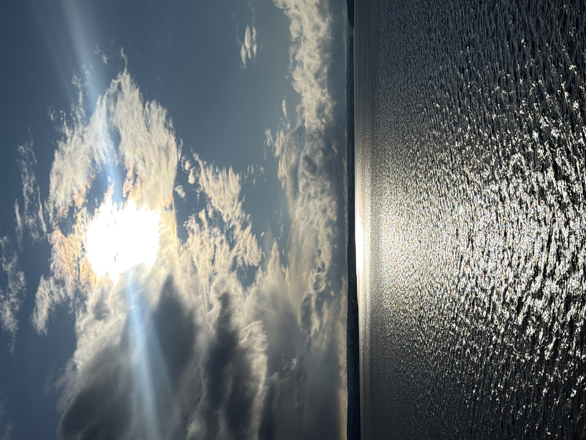 The image shows a view of the sky and water, possibly a lake or reservoir, under bright sunlight. The sun is shining through the clouds, creating rays of light. The water surface reflects the sunlight, giving it a shimmering appearance. The image appears to be rotated to the side. Given the context of Votkinsk, Russia, it is likely this is a view of the Votkinsk Reservoir. There are no people visible.