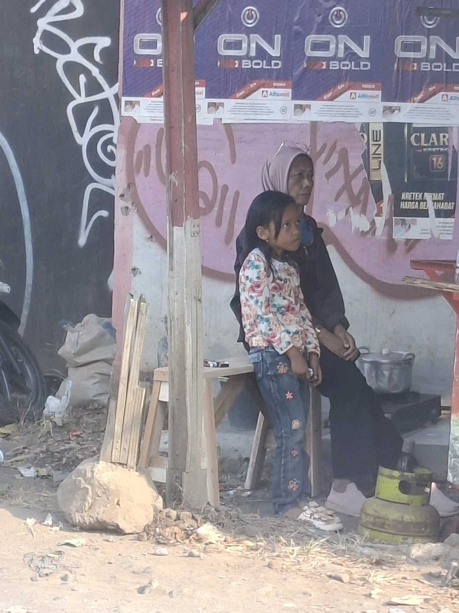 In Karangtengah, Indonesia, a young girl with dark hair is standing on the left, facing to the right and looking away from the camera. She is wearing a long-sleeved shirt with a floral pattern and blue jeans adorned with heart-shaped patches. To her right, an adult, appearing to be a woman wearing a light-colored hijab and dark clothing, is seated, facing towards the left and slightly away from the camera. Their hands are clasped.
Behind them, a rustic wooden structure, possibly a makeshift stall, provides shade. The background features a wall covered in peeling posters, some displaying advertisements with text in Indonesian and English, including "ON BOLD" and "CLAR". Graffiti in pink and white spray paint is visible on the wall.
The ground is a mix of dirt, debris, and scattered rocks. To the left, a large, rough-textured rock is prominent. In the lower right corner, a green gas cylinder rests near a stove. A metal pot sits on the stove. Scattered pieces of wood are leaning against the structure. The overall environment appears to be an outdoor, somewhat cluttered, urban setting.