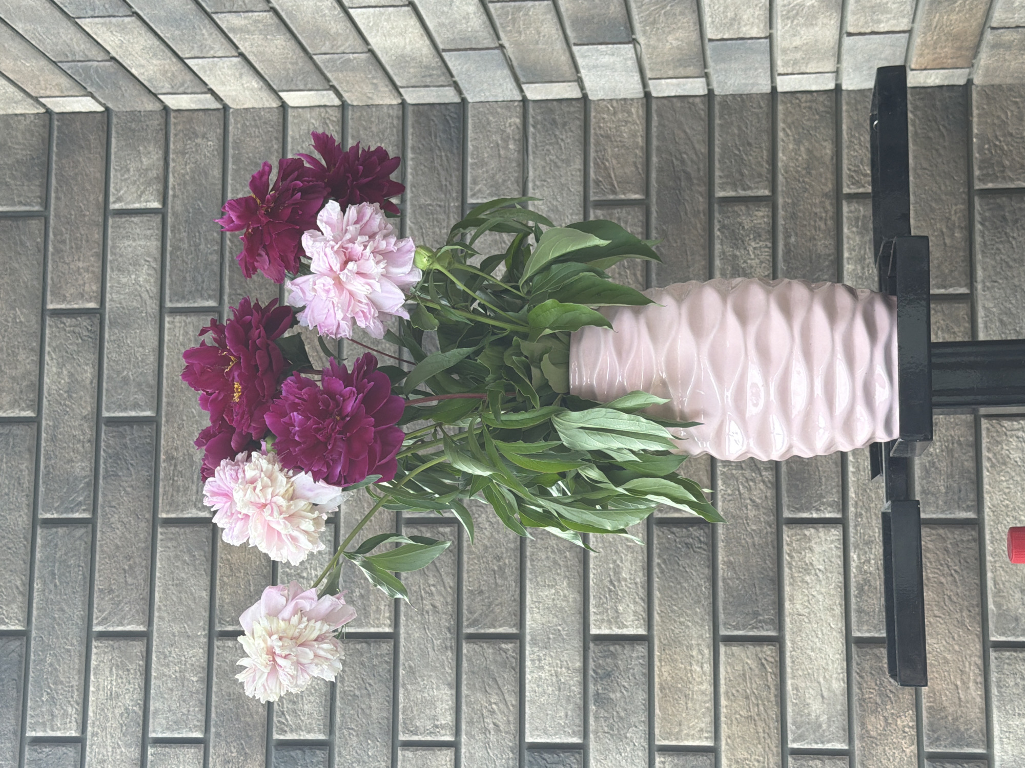 A bouquet of peonies in shades of pink and dark magenta is displayed in a pink wavy vase. The vase sits on a black metal stand in front of a textured brick wall. The wall features horizontal rows of greyish-brown bricks with variations in tone and a rough finish. The lighting suggests an outdoor setting, possibly a patio or balcony. No people are present in the image. The scene is static and appears to be a still life composition. The image was taken in Bila Tserkva, Ukraine.