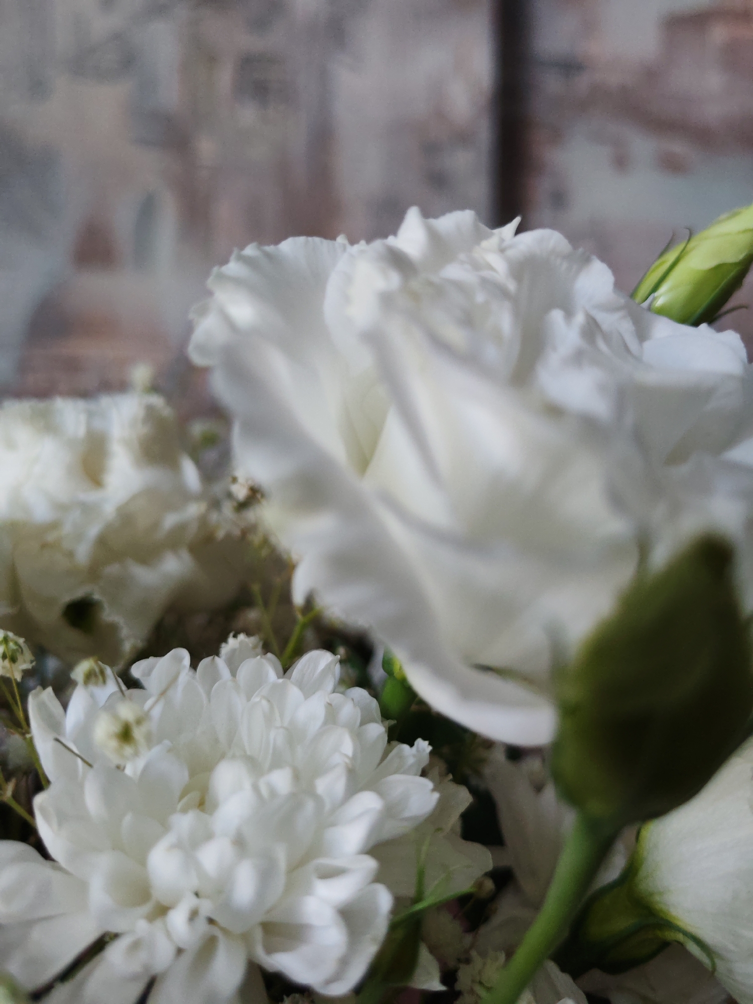 This is a close-up photograph of a bouquet of white flowers against a blurred, textured background. The flowers appear to be a mix of varieties, including what looks like a chrysanthemum with many delicate petals in the foreground, and larger, fuller blooms with layered petals in the background. There are also some small, delicate white florets and green buds or stems visible. The lighting highlights the soft textures and white hues of the flowers. The background is out of focus, showing a pattern that could be a wall covering or fabric with a muted, abstract design. There is no indication of the city of Yekaterinburg, Russia, or any specific location. The image depicts a still life arrangement of flowers.
