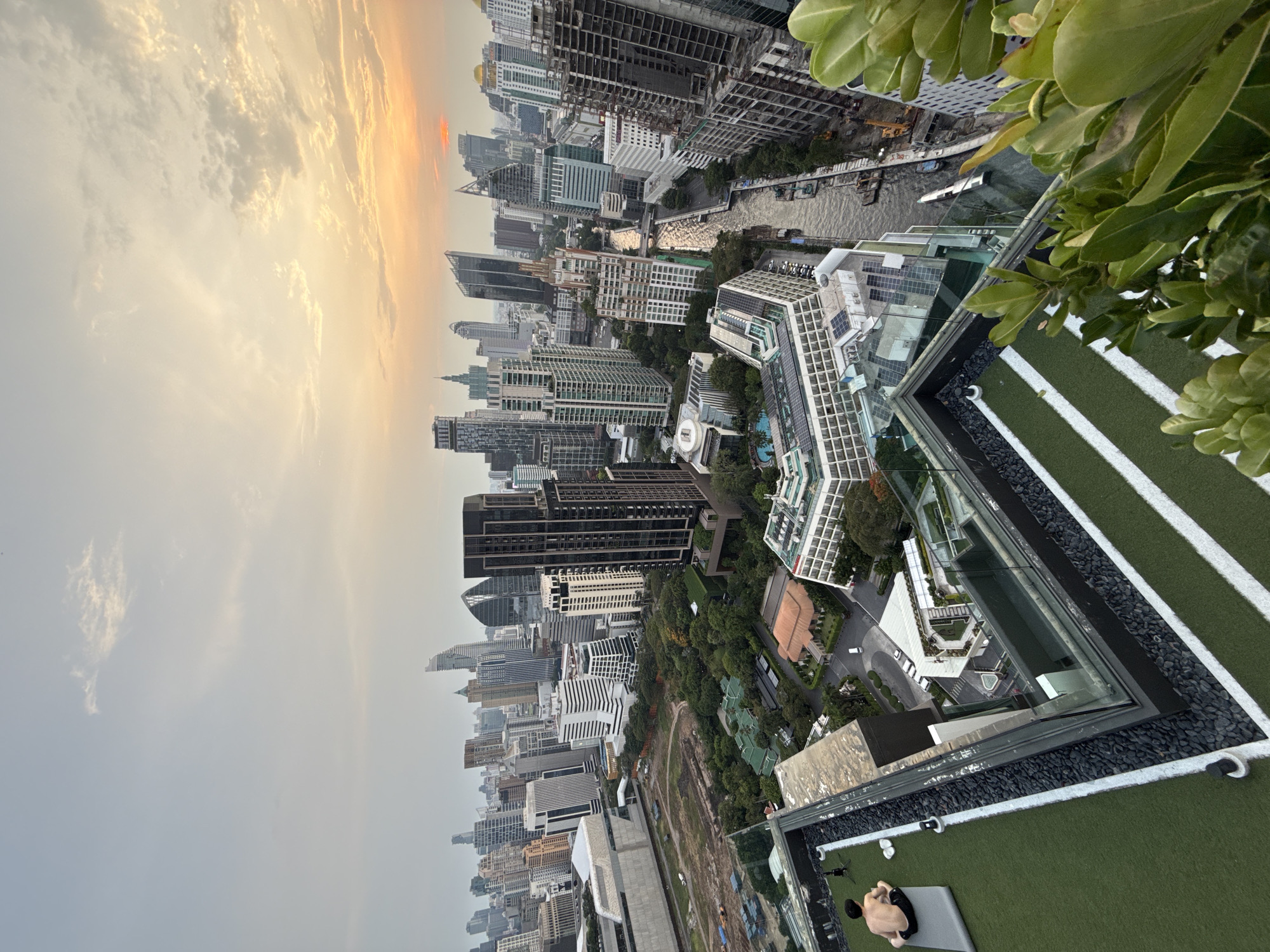 The image depicts a rooftop view of Bangkok, Thailand, at dusk. Buildings dominate the cityscape, with varying heights and architectural styles. A river or canal winds through the urban landscape. In the foreground, a rooftop area features artificial turf with white stripes and a glass barrier. A person is lying on a mat on the rooftop, appearing to be relaxing or sunbathing. The sky is overcast, with the sun setting on the horizon. Green foliage is visible in the right corner of the frame.