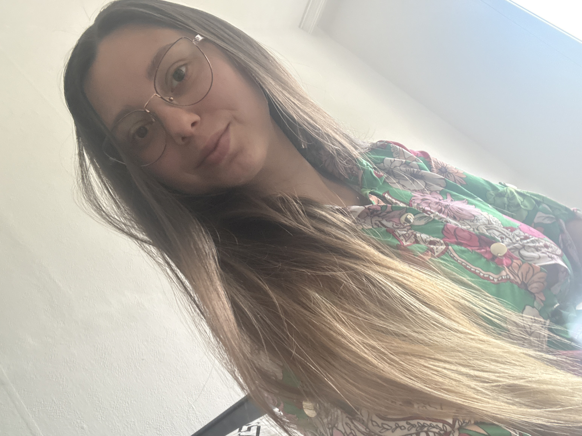A young woman with long, blonde-brown hair and glasses is captured in a selfie. She is wearing a green shirt with a floral pattern. The background is a plain white wall. The lighting suggests an indoor setting, possibly a room in a residential building. The angle of the photograph indicates it was taken from a low viewpoint. There is no identifiable information in the image to suggest a specific location within the city of Plovdiv, Bulgaria.