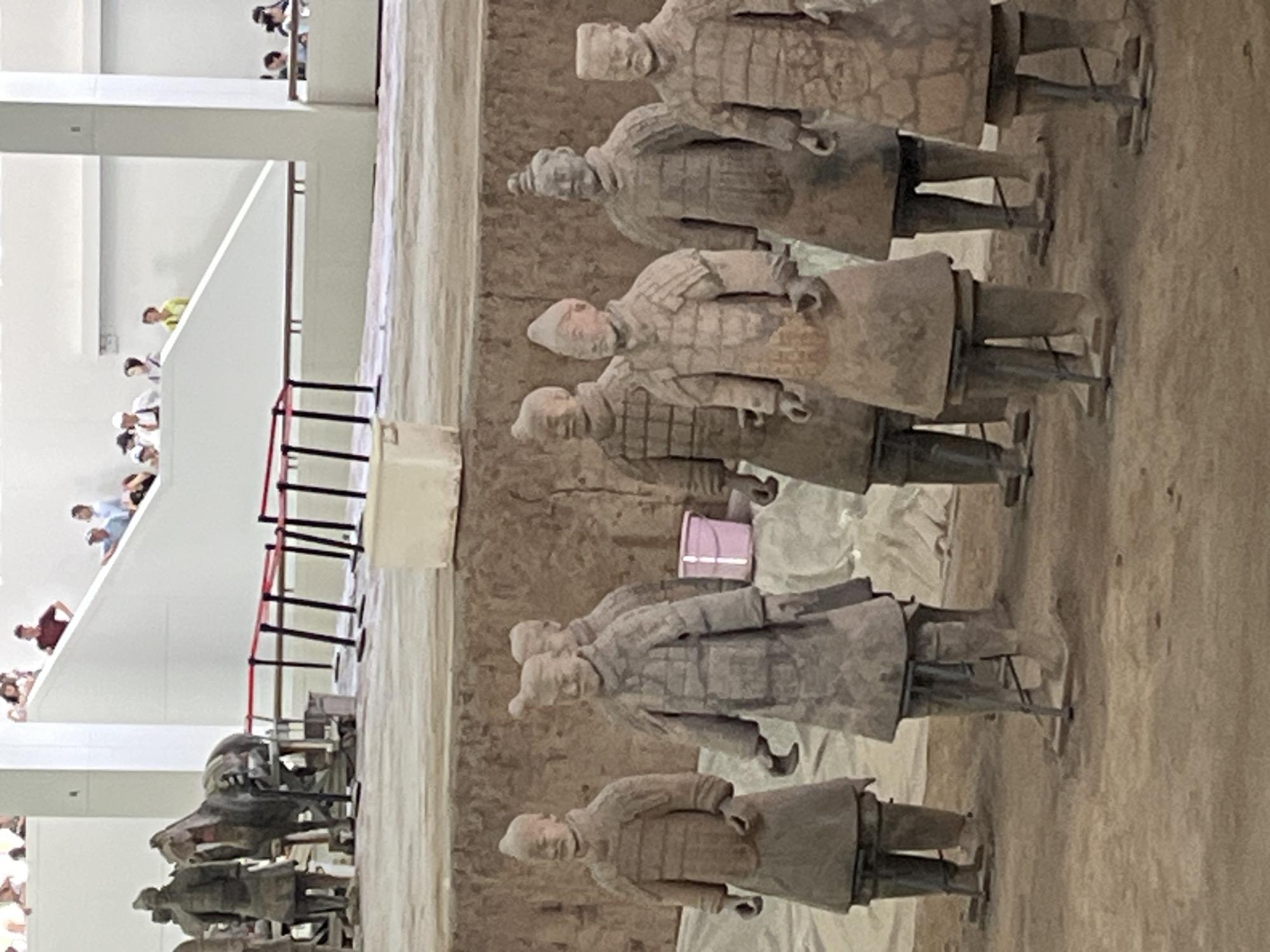 The image depicts the Terracotta Army, a collection of terracotta sculptures depicting the armies of Qin Shi Huang, the first Emperor of China. In the foreground, several life-sized terracotta warrior figures stand in formation, clad in ancient armor and positioned on square bases. Their faces are distinct, with individual hairstyles and expressions. Behind them, a row of terracotta horses and chariots are visible, further illustrating the grandeur of the army. The figures are arranged in a pit-like structure, characteristic of the archaeological site where they are displayed. In the background, a crowd of onlookers can be seen observing the exhibit from a raised walkway. The environment appears to be a museum or exhibition hall, with muted lighting and a backdrop of earth-toned walls, suggesting an archaeological preservation setting. The scene conveys a sense of historical significance and the immense scale of the ancient Chinese military.