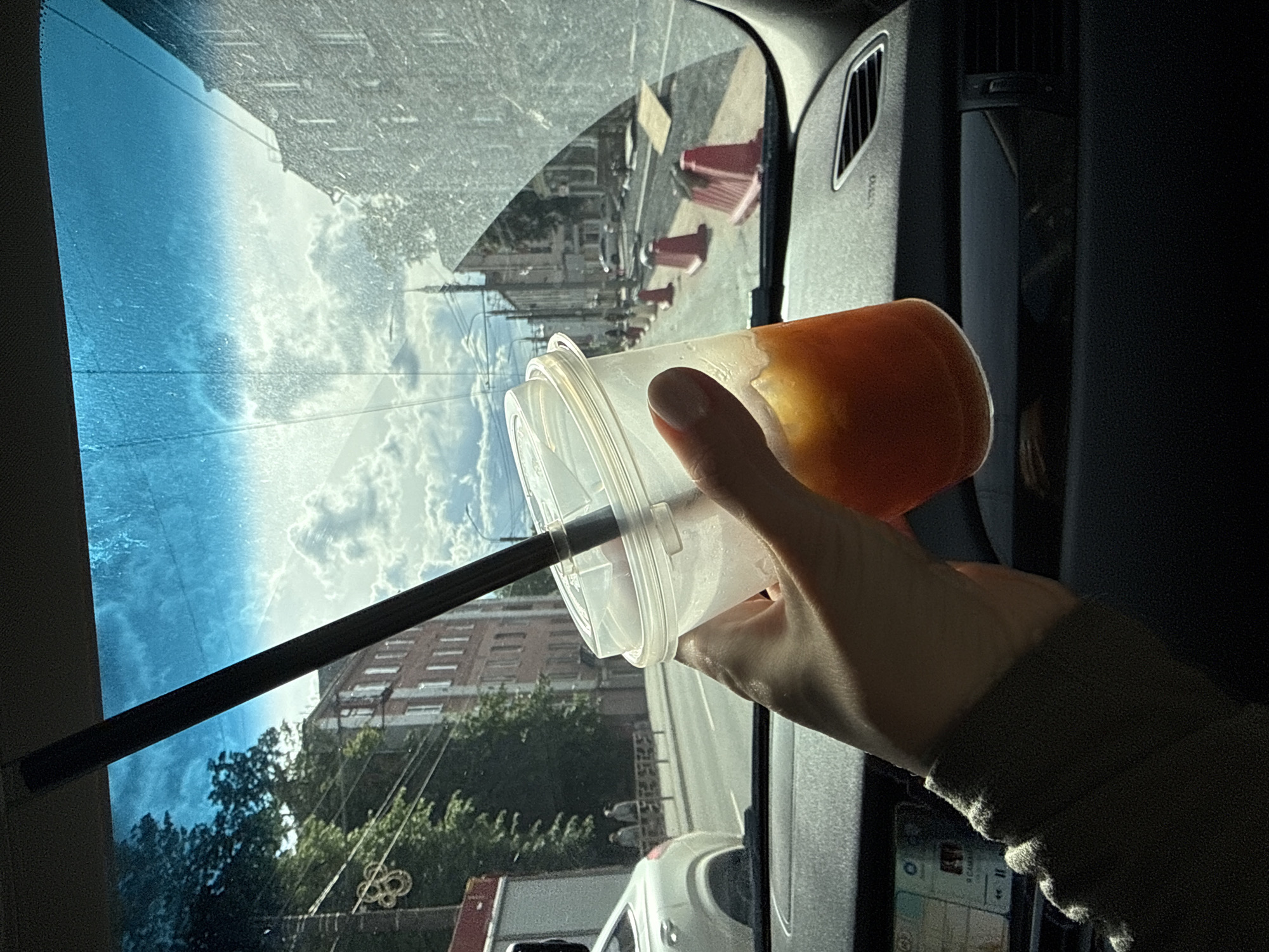 A hand holds a plastic cup with a straw in the front seat of a car. The cup contains an orange liquid. Outside, a city street with buildings and trees is visible through the windshield. Orange traffic cones line the road. The car is presumably in Izhevsk, Russia.