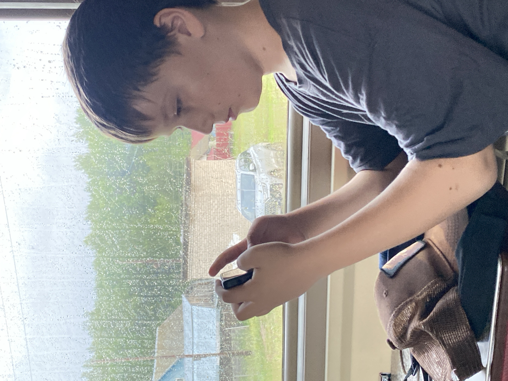 The media file shows a young person looking down at a phone, their hands holding the device. The person is wearing a dark grey t-shirt. To the left of the person is a window with raindrops on it, suggesting it is raining. Outside the window, blurred greenery and what appear to be buildings are visible. A brown baseball cap is draped over something behind the person. The lighting and rain on the window indicate it is likely daytime, possibly overcast due to the weather. The scene appears to be indoors, by a window, with a view of an outdoor environment. The person's focus on the phone suggests engagement with a digital activity. There is no visible text in the media file.