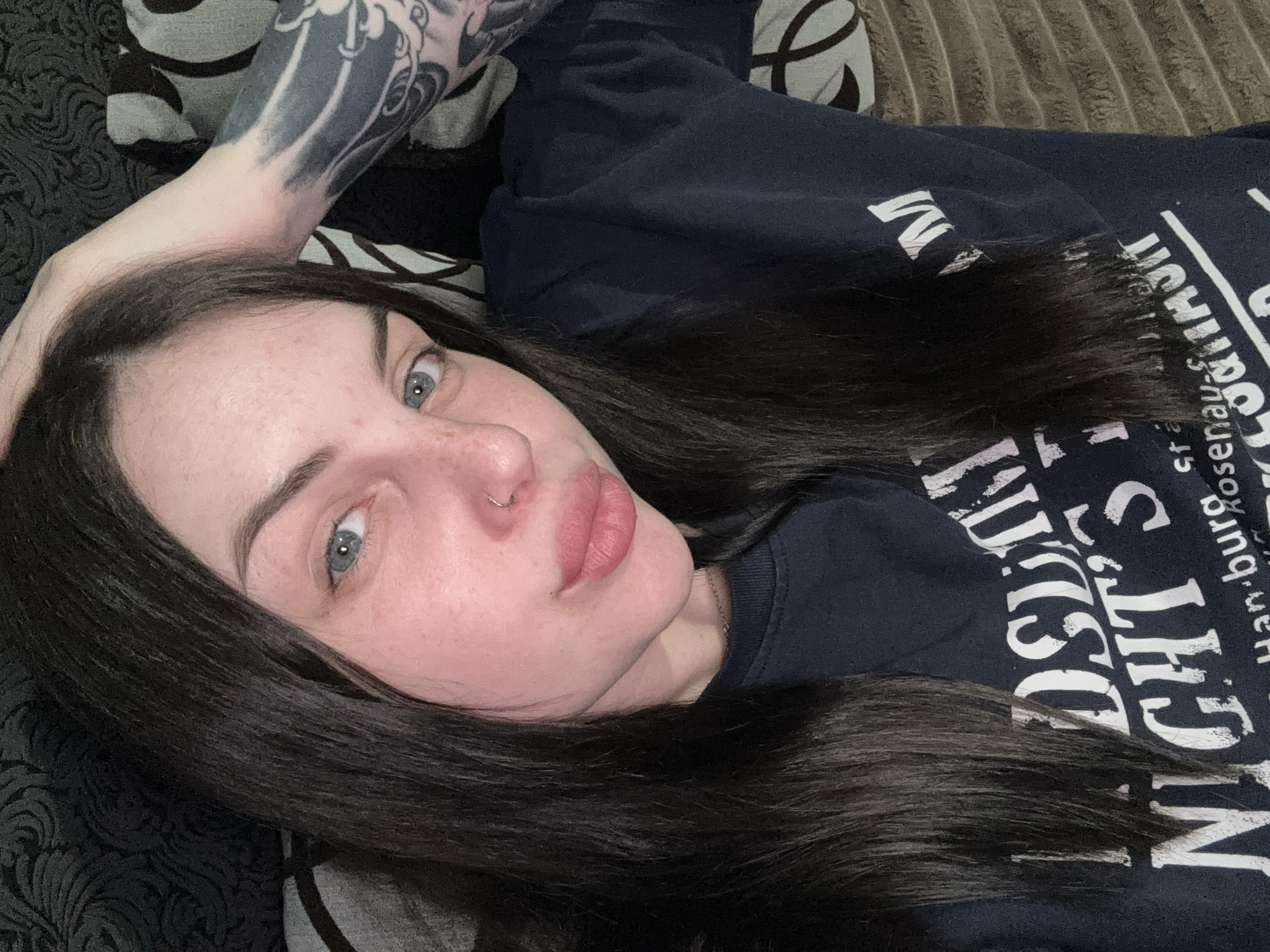 A woman with fair skin, dark hair, and blue eyes is lying down. She has a nose piercing and some freckles on her face. Her right arm is bent with her hand near her head, revealing a tattoo on her forearm. She is wearing a dark-colored shirt with white text on it. She is lying on a dark patterned fabric. The location is not provided in the image.