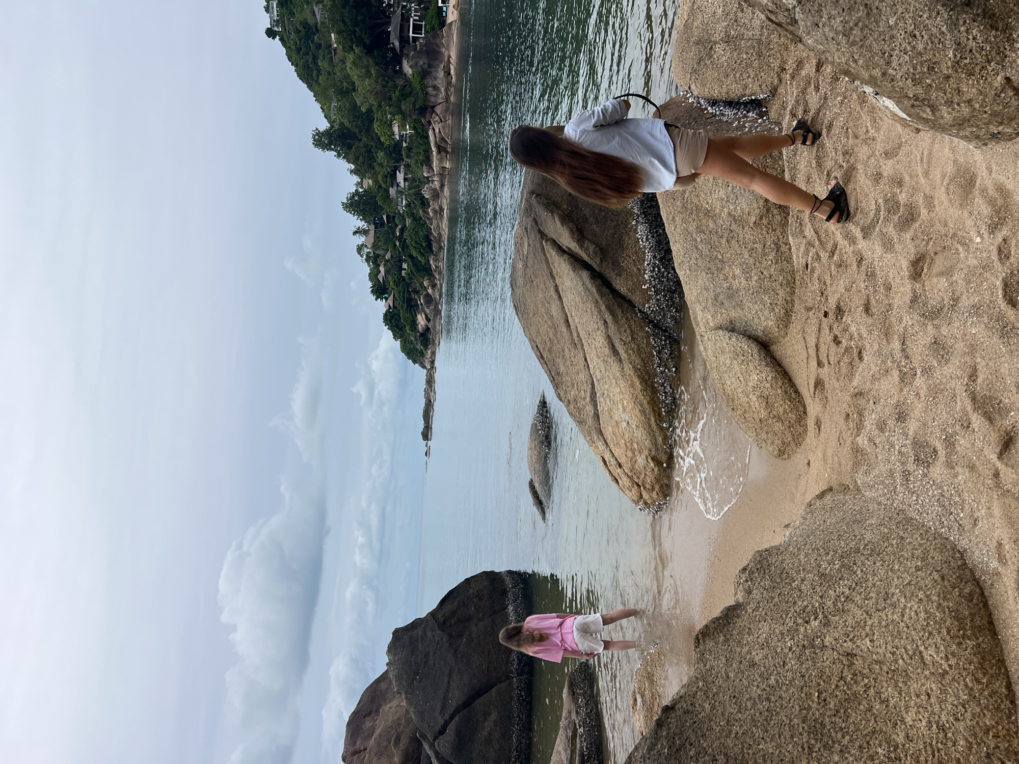 The image depicts a cloudy day at a rocky beach in Baan Chaweng Noi, Thailand. Two individuals, both appearing to be young females, are present. One is standing in the shallow water near a large dark boulder, wearing a pink t-shirt and white shorts. The other is walking on the sandy shore away from the water, wearing a light blue button-up shirt and beige shorts, with dark sandals. The background shows a calm ocean with a distant green, hilly coastline dotted with buildings and trees. Large, weathered rocks are scattered along the shore.