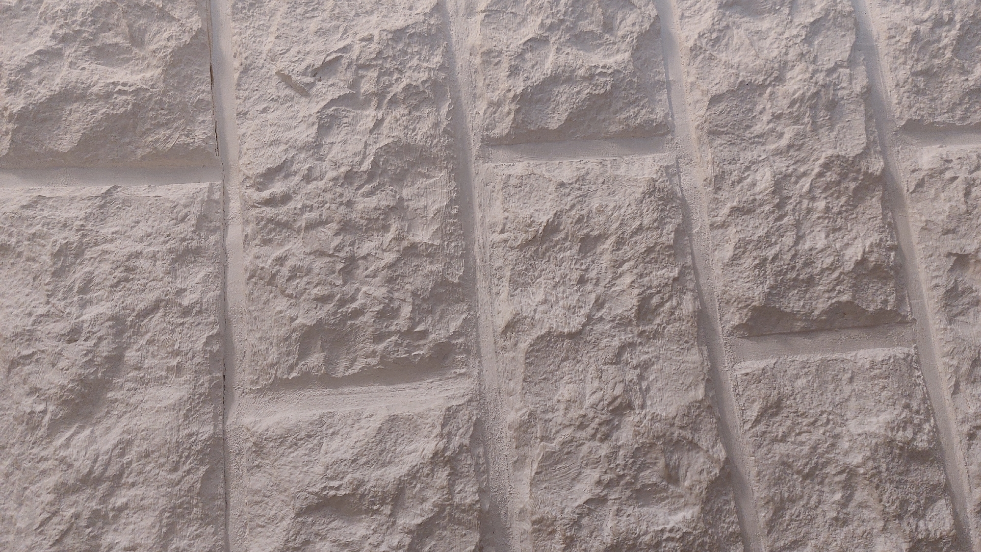 The image shows a close-up, textured view of a light-colored, rough-hewn stone wall. The stones are rectangular and laid in a staggered pattern, with visible mortar lines between them. The surface of each stone is uneven, with variations in depth and texture, suggesting natural stone or a realistic imitation. The lighting casts subtle shadows, highlighting the three-dimensional quality of the stone work. The background is entirely composed of this stone wall, and there are no people, animals, or other objects visible. The scene is static and appears to be an architectural detail. There is no information available to pinpoint its specific location within the city of Bad Laasphe, Germany.