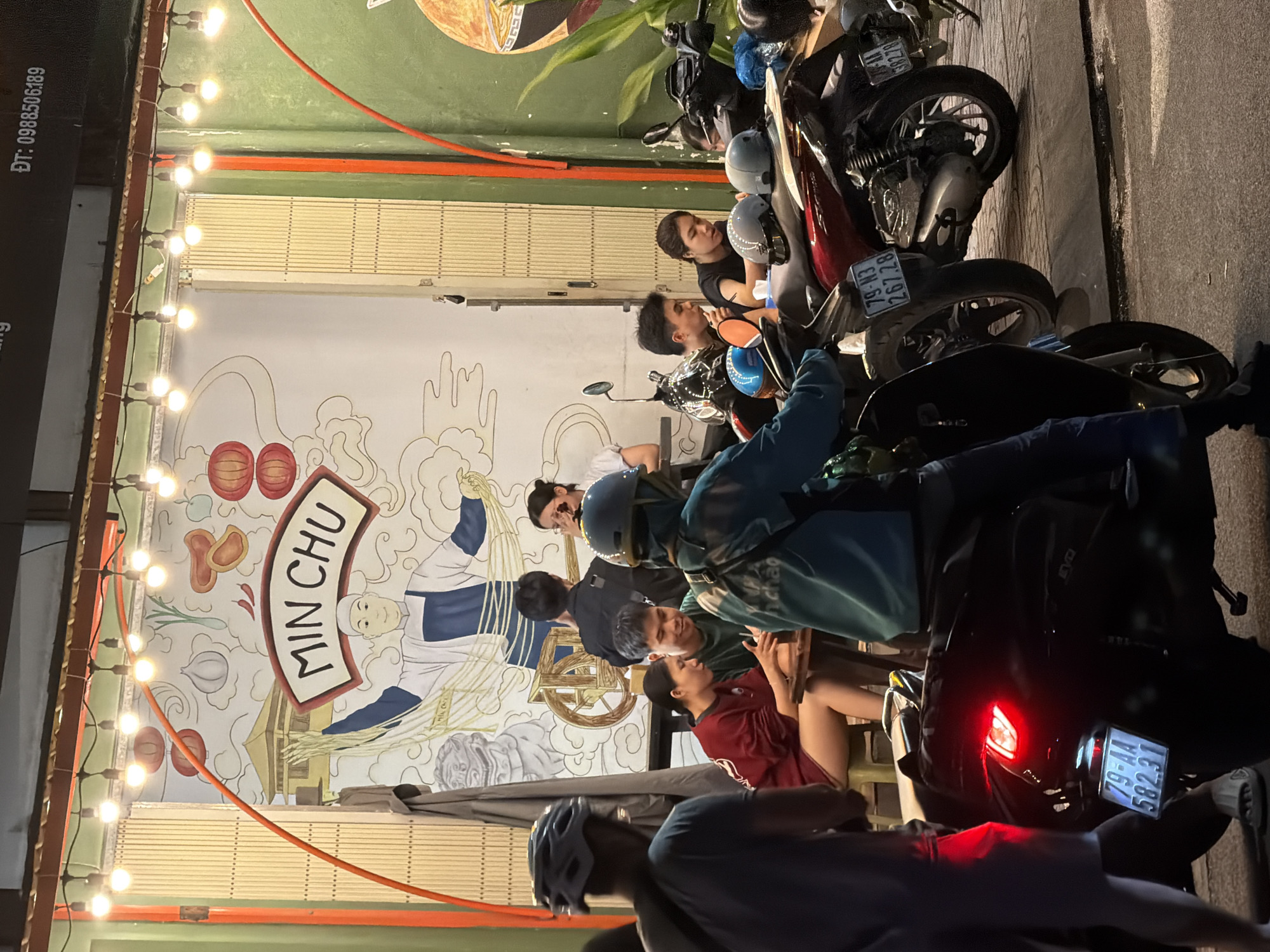 The image depicts a street scene at night in Nha Trang, Vietnam, outside a restaurant called "MIN CHU". A mural of a chef with the restaurant's name is visible on the wall behind a group of people seated at a table. Several motorbikes are parked on the sidewalk, some with helmets on them. The scene is illuminated by string lights and the red taillight of a motorbike in the foreground. A delivery driver, wearing a blue uniform and helmet, is positioned on a motorbike, facing away from the camera. The atmosphere appears casual and social, with people engaged in conversation and dining.