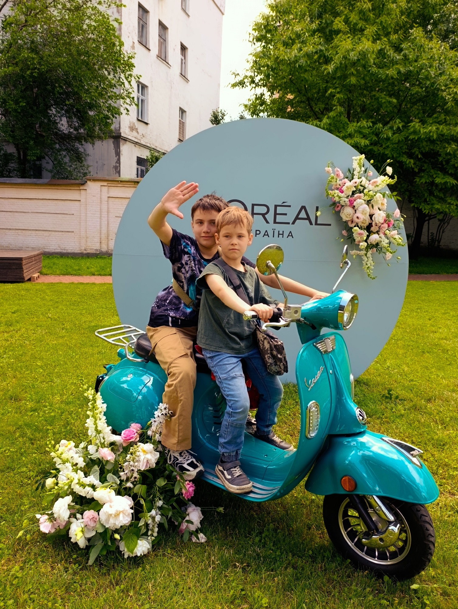 Two boys are sitting on a teal-colored scooter in a grassy outdoor area. The older boy, in the back, is wearing a dark t-shirt with a graphic print and tan cargo pants. He is waving at the camera. The younger boy, in the front, is wearing a dark t-shirt and blue jeans. He is looking at the camera with a neutral expression.
The scooter is decorated with a large bouquet of white and pink flowers at the front, cascading onto the grass. A similar floral arrangement is attached to a large, light blue circular backdrop behind them. The backdrop has text on it, including "L'ORÉAL" and "УКРАЇНА" (Ukraine in Cyrillic).
In the background, a white building with multiple windows and a tree with green foliage are visible. The ground is covered in green grass, suggesting a park or outdoor event space. The lighting indicates it is daytime, likely overcast or with diffused sunlight. The overall mood seems to be playful and celebratory, possibly for a promotional event. The location is identified as Kyiv, Ukraine.