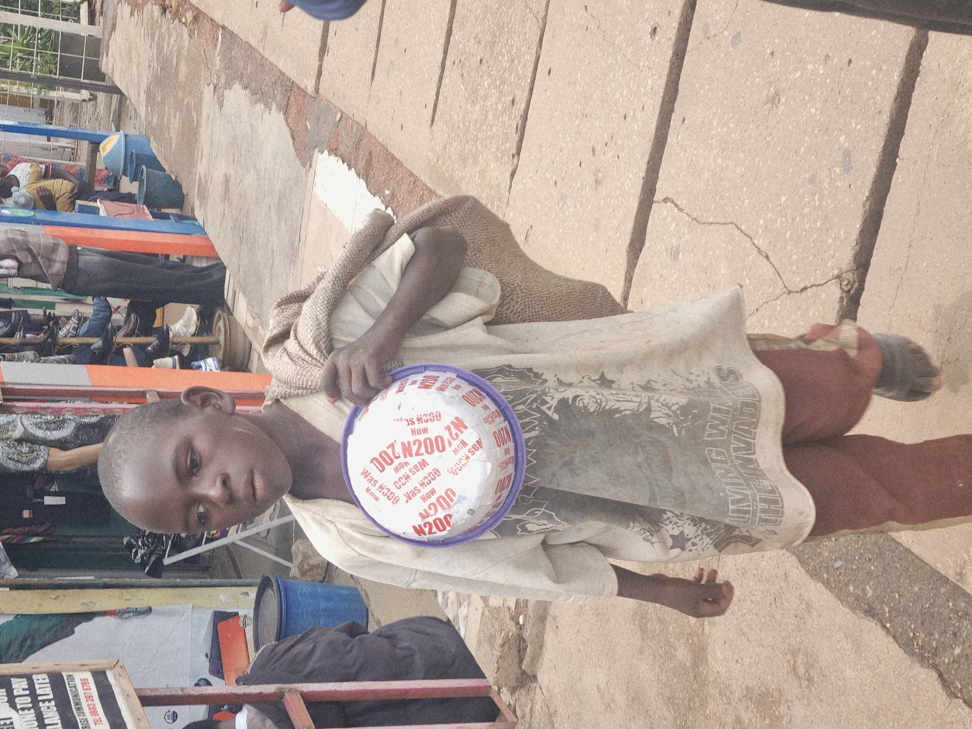 This image, taken in Jos, Nigeria, captures a young boy holding a plastic bowl filled with what appears to be coins. He is dressed in a light-colored t-shirt with a graphic design and a darker colored pair of pants. A woven sack is draped over his shoulders and torso. The boy is positioned on a concrete walkway, with a wall or building visible in the background. The scene suggests a street or market environment, possibly indicating the boy is collecting alms or selling something. The lighting implies it is daytime. Visible text on the bowl includes "N200" and "Was #300," suggesting a price change or a sale. Additional text on a sign in the lower left corner mentions "ONE TO PAY," "LANCE LATER," and "BISE COMMUNICATION TEL: 0803 397 8789," further indicating a commercial or service-oriented location.