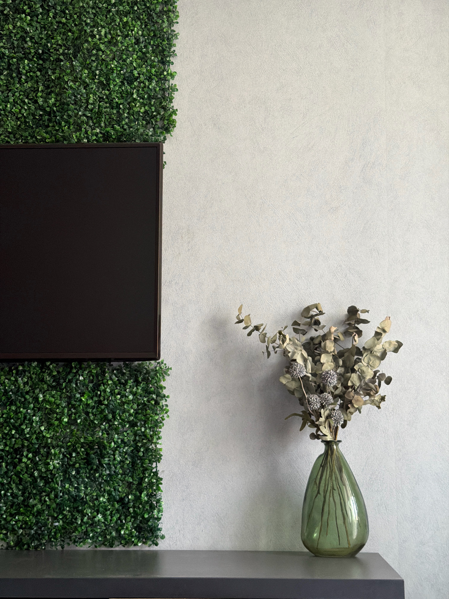 The image shows a portion of a room with a dark, flat-screen television mounted on a wall. The wall to the left of the television is covered in a dense, green artificial hedge, which also extends below the television. To the right of the television, the wall is a textured, light gray. On a dark gray shelf or table in the foreground, a green glass vase holds a bouquet of dried eucalyptus branches and small, grey, spherical dried flowers. The scene appears to be indoors, in a modern setting. There is no indication of the city of Orsha, Belarus.