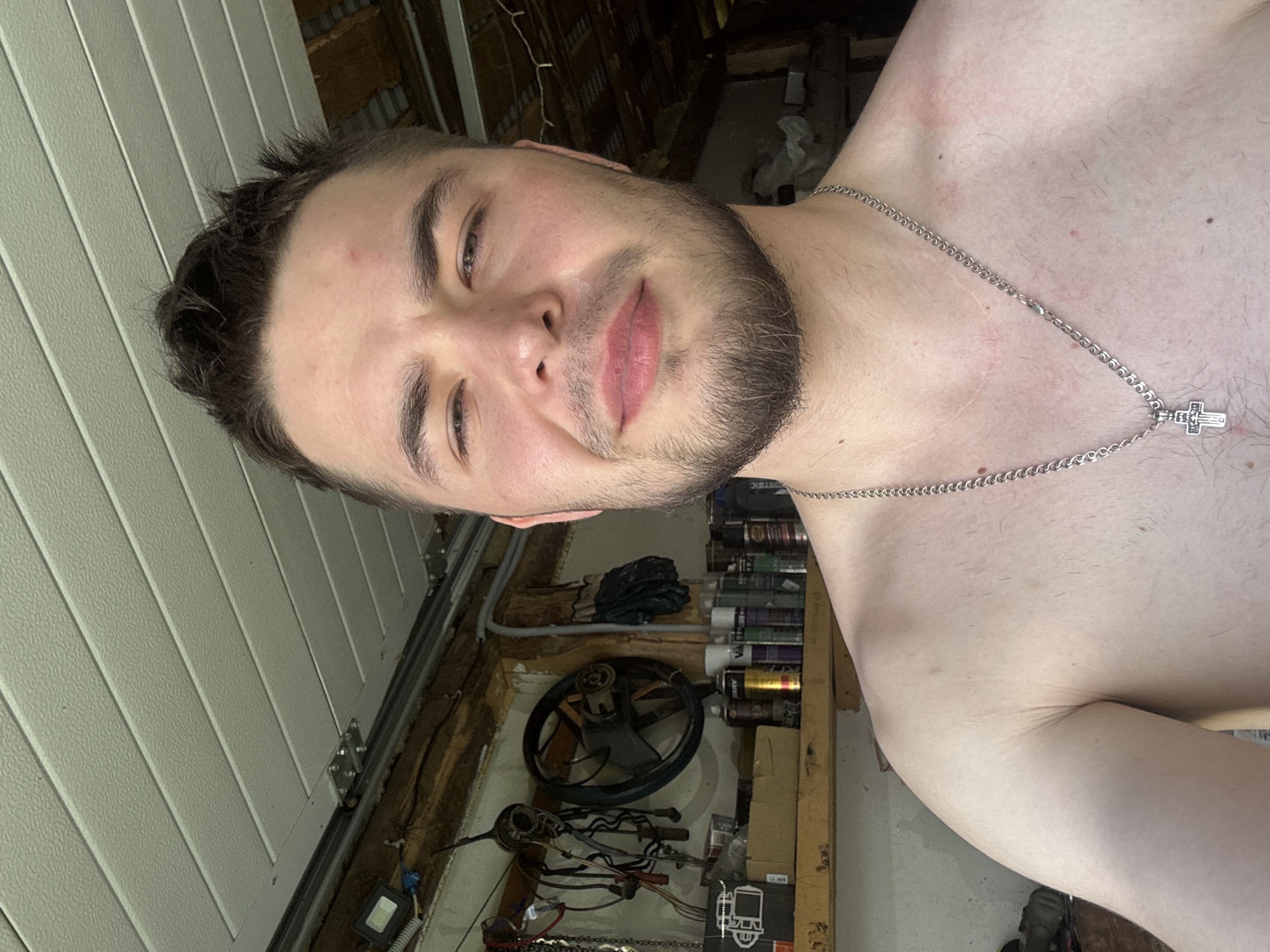 A medium shot shows a shirtless young man with light skin and brown hair, facing the camera. He has a short beard and is wearing a silver chain with a cross pendant. The background appears to be a cluttered garage or workshop with shelves holding various items, including spray cans, a steering wheel, and electrical wires. The lighting suggests an indoor setting, possibly with natural light coming from above. There are some red marks on the man's upper chest and shoulder. The overall impression is a casual, unposed moment captured in a utilitarian space.