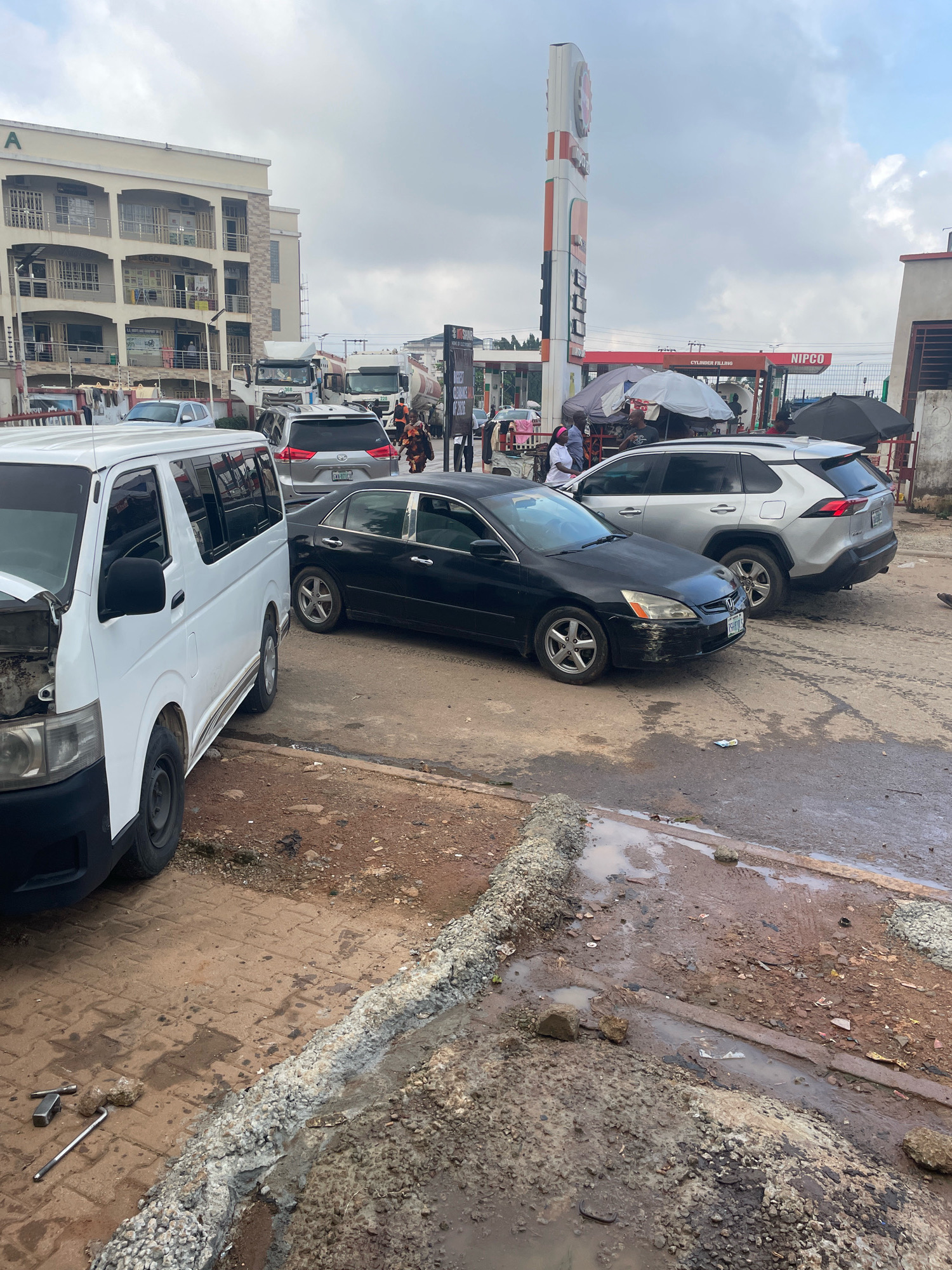 In Abuja, Nigeria, a street scene unfolds under an overcast sky, featuring a collection of vehicles and a multi-story building. In the foreground, a white van, possibly a commercial transport vehicle, is parked with its hood open, suggesting mechanical attention. Beside it, a black sedan is parked, its front bumper showing signs of damage. Further back, a silver SUV is also present.
The environment appears to be a mixed-use area, with a prominent NIPCO gas station and its tall sign dominating the background. Tents and makeshift stalls are set up near the gas station, indicating commercial activity. The ground is a mix of dirt, gravel, and some wet patches, hinting at recent rainfall or poor drainage. People are visible in the scene, some near the vehicles, others at the stalls or the gas station, engaged in various activities. The overall impression is one of a busy, functional, and perhaps somewhat unkempt urban environment.