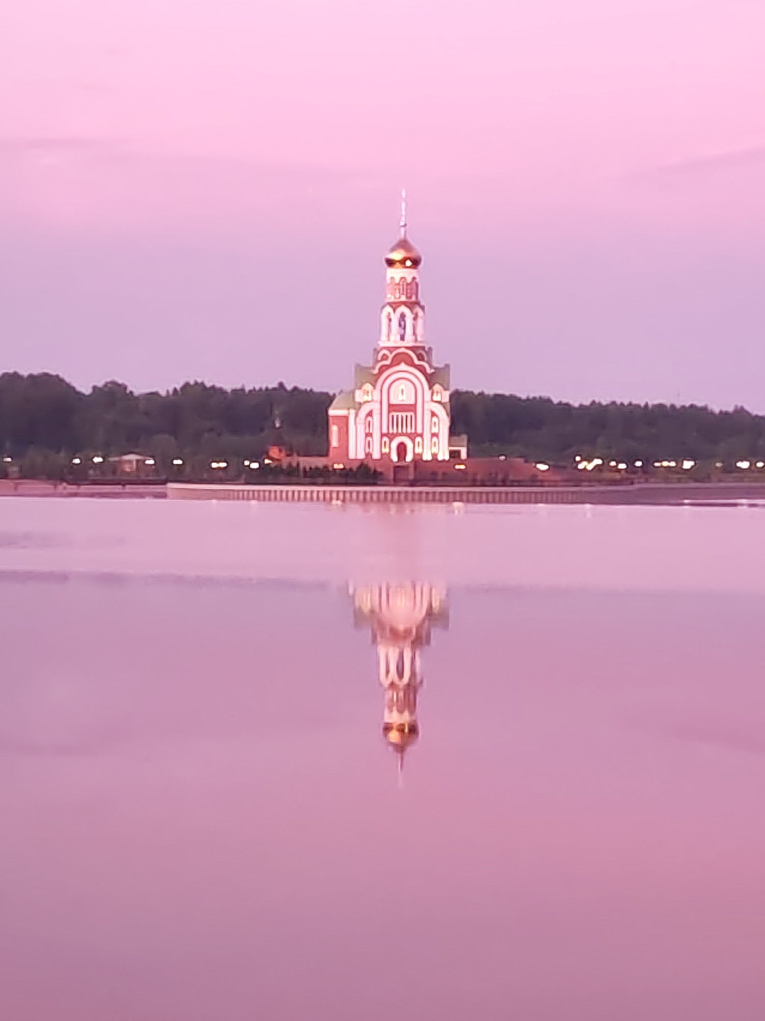 The image depicts the Church of the Assumption of the Blessed Virgin Mary, a prominent Orthodox church located in the city of Sverdlovsk, Russia. The church is characterized by its white walls adorned with red decorative elements and a tall, slender bell tower topped with a golden dome. It is situated on the bank of a body of water, likely a river or lake, which reflects the structure and the soft, pinkish hues of the sky during what appears to be either dawn or dusk. The background features a line of dark green trees, and along the waterfront, there are subtle lights that suggest buildings or a promenade. The water's surface is calm, creating a clear reflection of the church, enhancing the serene and picturesque quality of the scene. There are no people visible in the image.