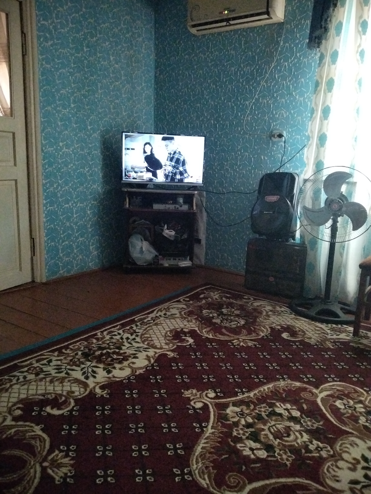 In a corner of a room in Kogon City, Uzbekistan, a television displays a couple in an indoor scene. The television is placed on a dark wood entertainment stand, which also holds various items on its shelves. To the right of the television, a tall, black oscillating fan with three blades stands on a pedestal, its base on a dark patterned rug. Behind the fan, a light-colored curtain with a floral pattern is visible. Above the television, an air conditioning unit is mounted high on the wall, which is covered in a light blue textured wallpaper. A white door is partially visible on the left side of the image. The floor is made of wooden planks, and the foreground is dominated by a large, ornate rug with a dark red base and intricate floral patterns in lighter shades.