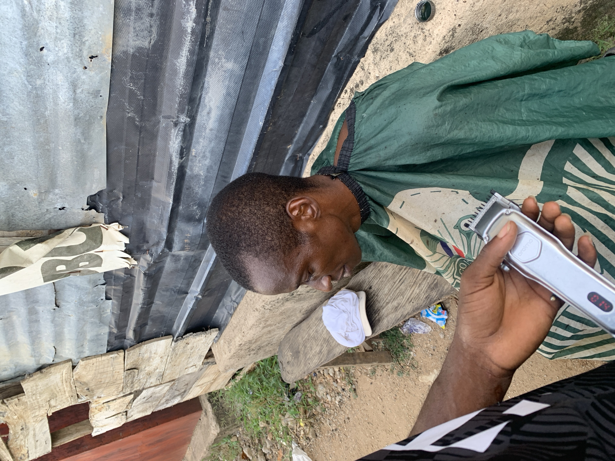 The image depicts a man receiving a haircut outdoors in Zango-ndaji, Nigeria. The man is sitting on a weathered wooden plank with a white cloth beneath his head, draped in a green and white patterned barber cape. A person, presumably the barber, holds a silver electric clipper. The background includes corrugated metal and makeshift wooden structures. Trash litters the ground.