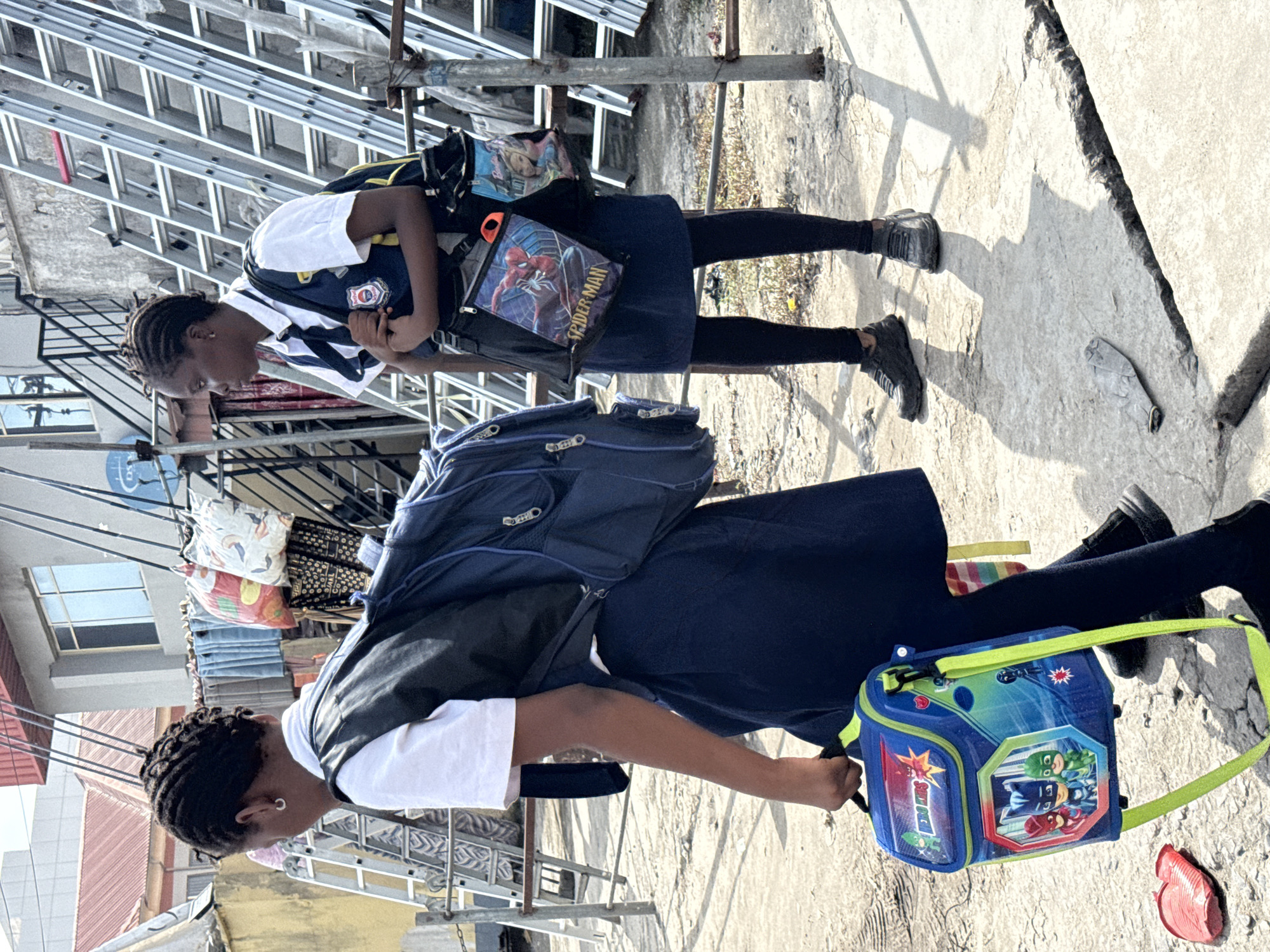 Two young girls are positioned on a concrete surface. The girl in the background has cornrows and is wearing a white shirt with a blue and white striped collar, a navy skirt, black leggings, and dark shoes. She carries a backpack and a Spider-Man-themed bag. The girl in the foreground also has cornrows and is wearing a white shirt, navy skirt, black leggings, dark shoes and carrying a backpack and a PJ Masks-themed lunchbox. A ladder leans against the structure in the background. The concrete surface is cracked. In the background, there is laundry hanging outside, and a building can be seen. The image appears to be taken in Ado, Nigeria.