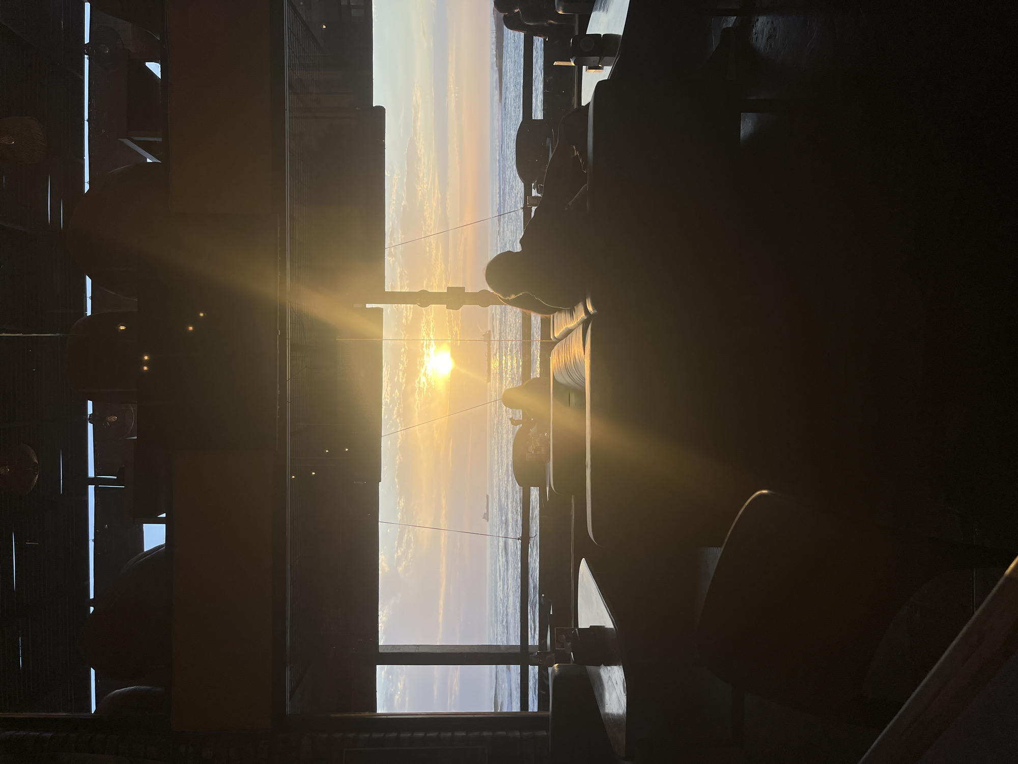 The image shows a sunset view through a window, likely a window facing west over the Pacific Ocean in Arica, Chile. The bright sun is visible on the horizon, with the ocean and sky filled with light clouds. Silhouettes of indoor furniture and a person are in the foreground, obscuring part of the view. A window frame is visible on the left. The image is rotated 90 degrees counterclockwise.