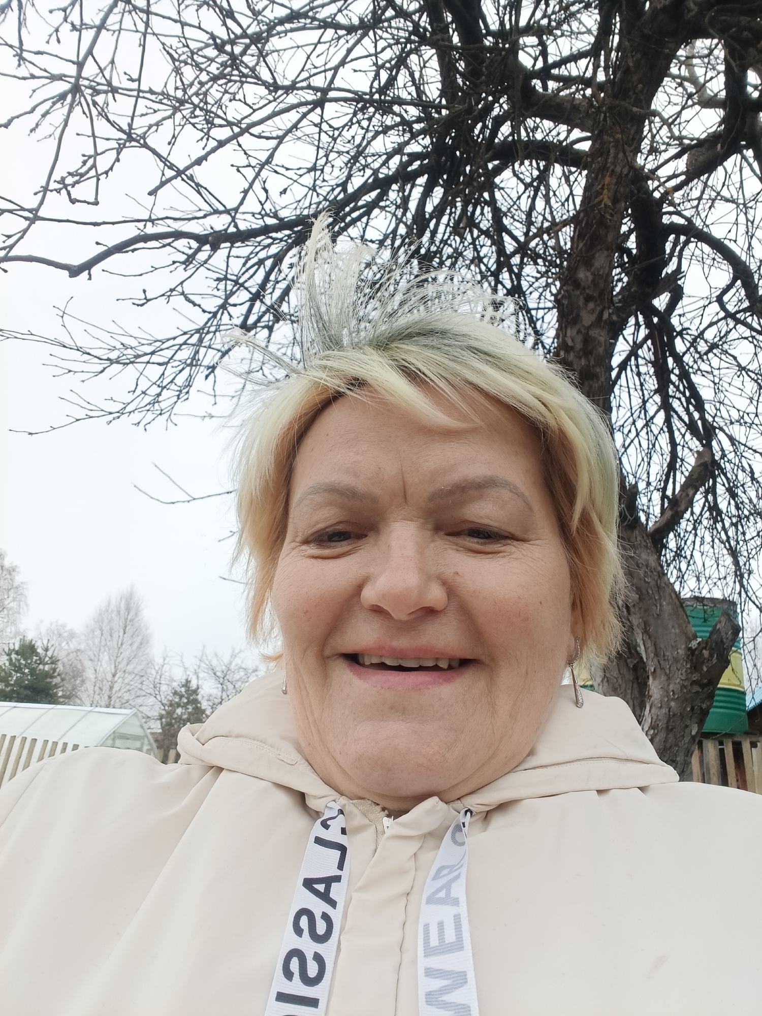 A close-up selfie depicts a smiling older woman with short, light blonde hair in an outdoor setting. She is wearing a light beige hooded jacket with the word "CLASSIC" repeatedly printed on the drawstrings.
Behind her, a large tree with bare branches dominates the background. To the left, other smaller trees and a greenhouse-like structure are visible. A green and brown wooden structure is visible to the right of the tree.
Given the prompt's mention of Красный Маяк, Russia, it is reasonable to assume the image was taken there. However, without explicit visual confirmation, the location within the city is unknown.