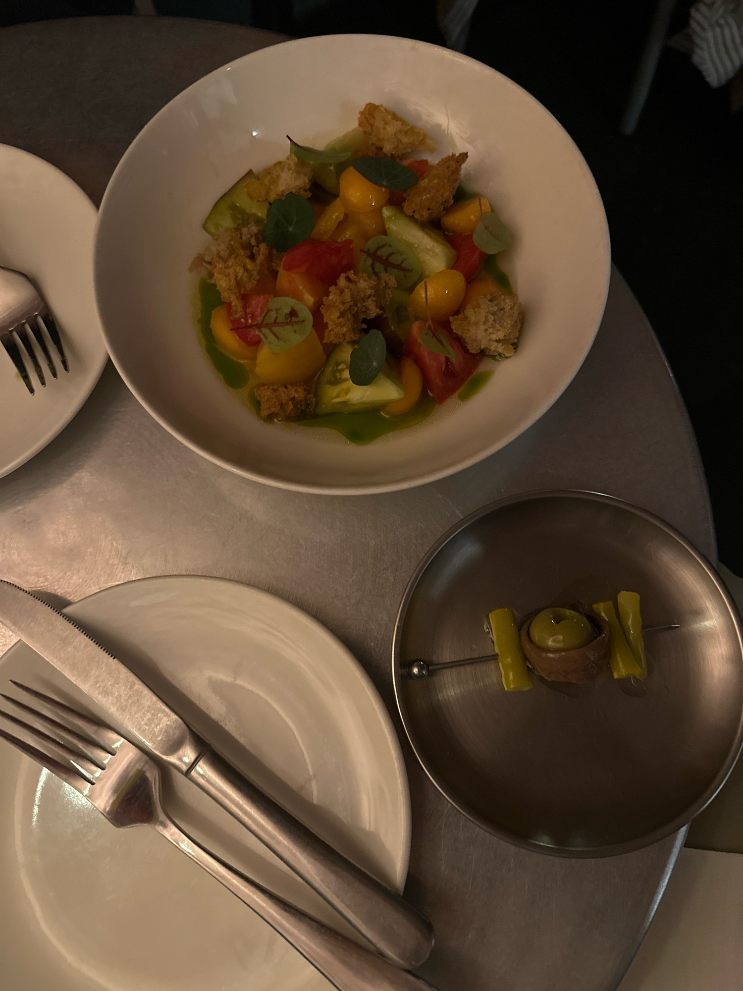The image depicts a close-up, overhead view of a dining table setting. In the upper portion of the frame, a white bowl contains a salad with diced tomatoes of various colors, green vegetables, and what appear to be croutons or crispy bread pieces. The salad is dressed with a green sauce and garnished with small green leaves. To the left of the bowl is a white plate with a fork resting on it. Below this, a larger white plate is visible with a knife and fork arranged on it. In the lower right quadrant, a small, dark metallic dish holds a skewer with an olive, an anchovy, and two pieces of green pepper. The table surface appears to be metallic and reflective. The lighting suggests an indoor setting, likely a restaurant. There are no people visible in the image. The location is not specified within the image or its context, and therefore cannot be identified as Madrid, Spain.