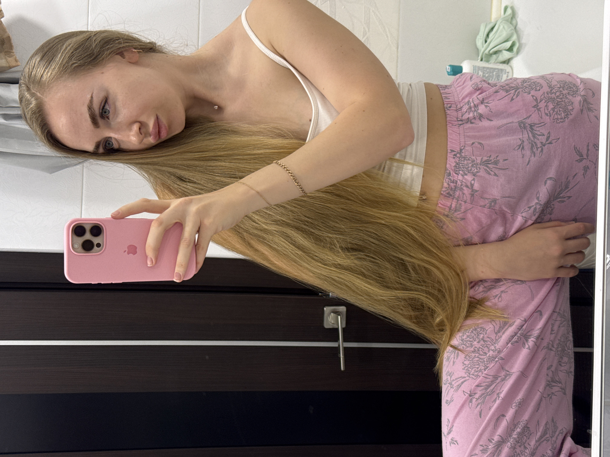 A blonde woman is holding a pink iPhone in a bathroom, seemingly taking a selfie. She is wearing a white tank top and pink floral patterned pajama pants. The background includes a dark brown cabinet with a silver handle and a white tiled wall with toiletries in the upper right corner. The photo appears to be taken indoors, and given the limited visual information, there are insufficient details to determine if the photo was taken in Petropavlovsk-Kamchatsky, Russia.