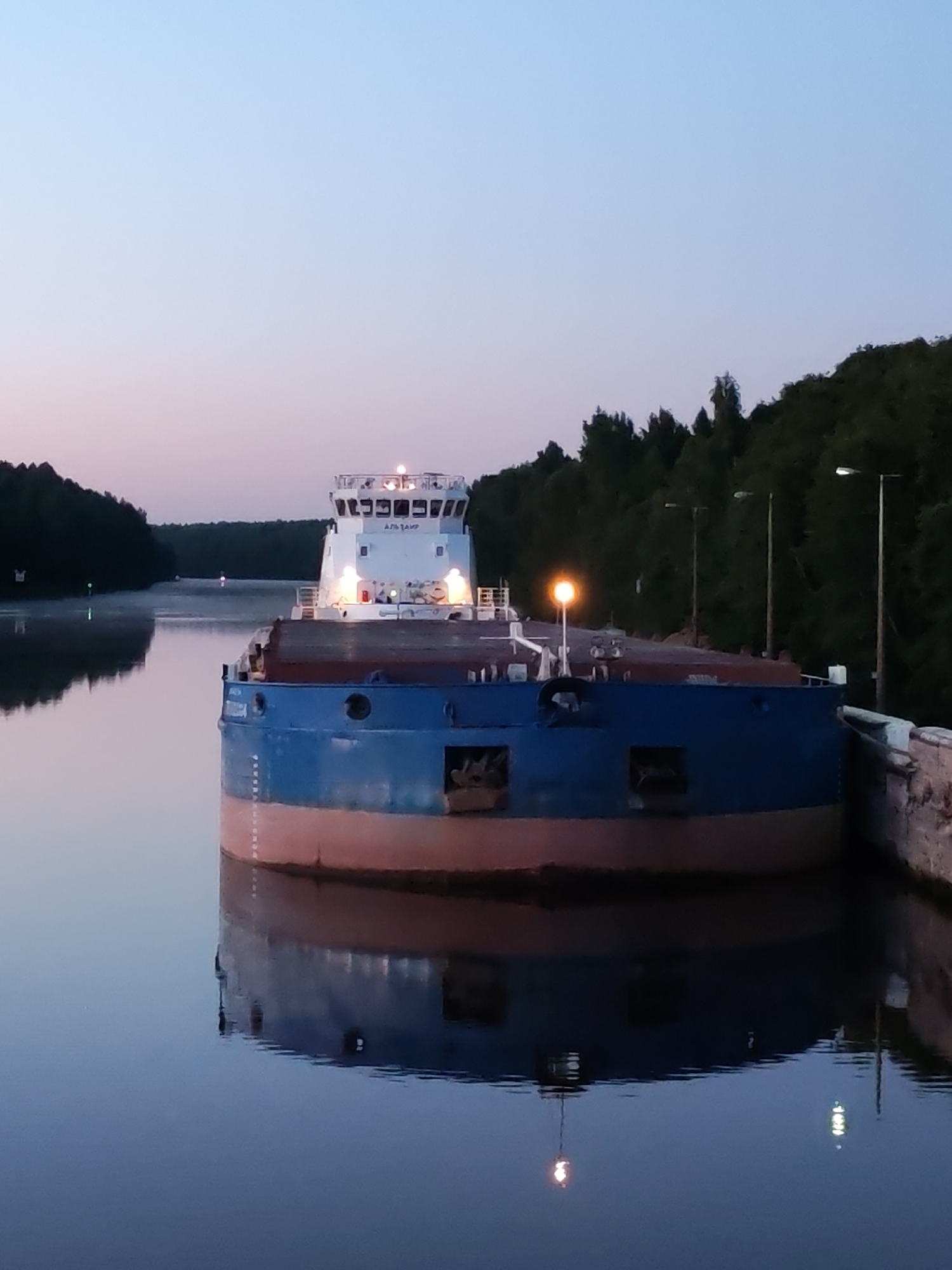 The image depicts a cargo barge docked at a canal in Devyatiny, Russia. The barge, painted blue and red, has a white superstructure with illuminated windows and a light on its front. The sky is a gradient of soft blue and pink, suggesting either dawn or dusk. The water is calm and reflects the barge and the surrounding trees, which form a dark silhouette along the canal banks. Several lampposts are visible along the canal, also reflected in the water. The scene is serene and still, with no people visible. The overall atmosphere is peaceful and tranquil, highlighting the quiet beauty of the Russian landscape at twilight.