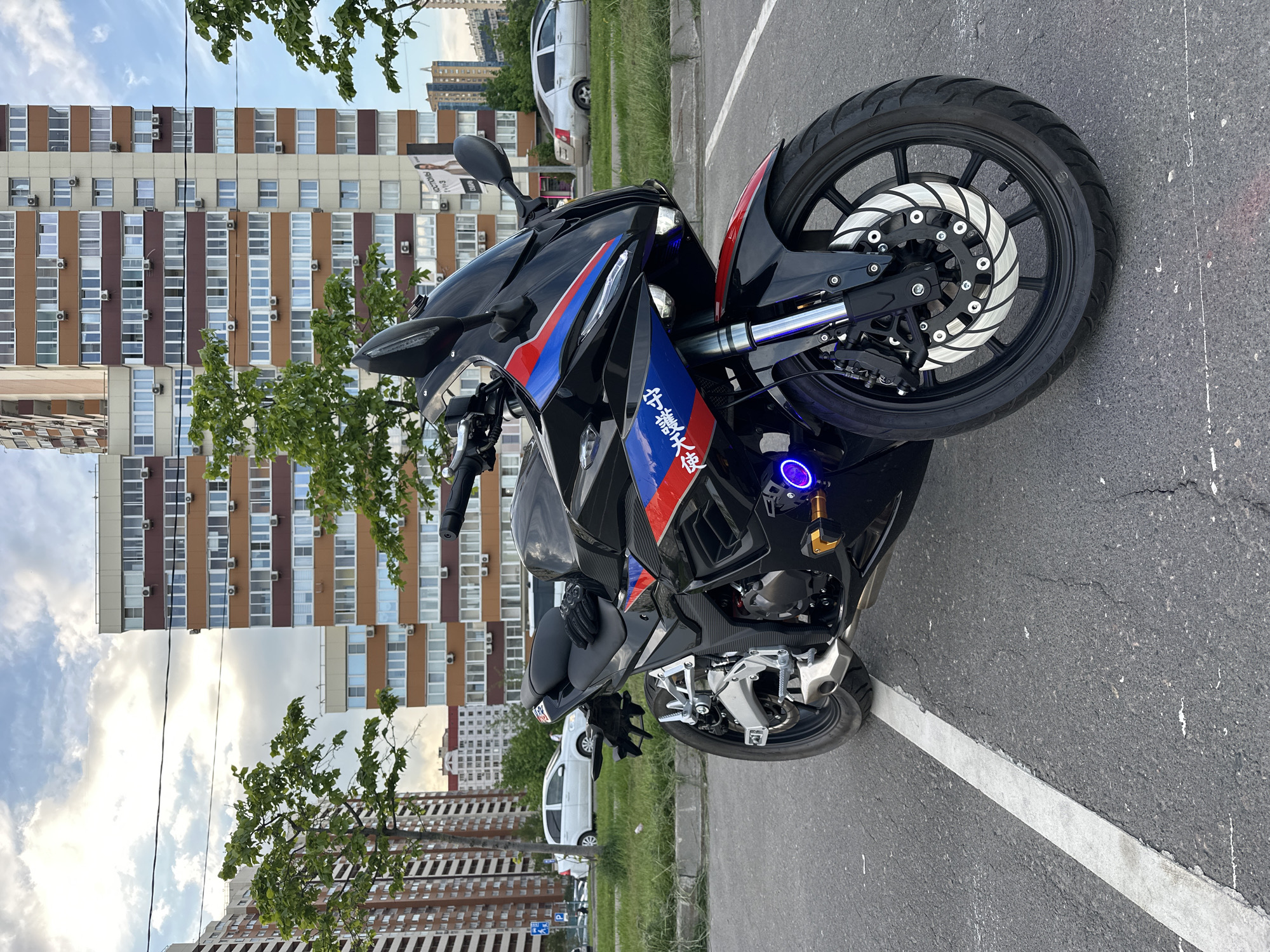 A black and blue BMW motorcycle is parked in a parking space in Moscow, Russia. The motorcycle has red accents and custom lettering on the side. A pair of black gloves are on the motorcycle seat. Tall, multi-story apartment buildings can be seen in the background, along with parked cars. The sky is partly cloudy. The motorcycle is parked on asphalt, delineated by a white parking line.
