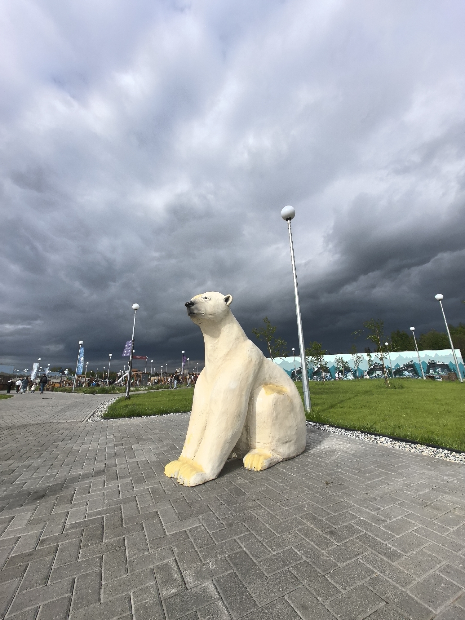 The image depicts a concrete polar bear statue in Perm, Russia. The statue is seated on a paved walkway constructed of gray rectangular bricks. The sky above is overcast with dark storm clouds. In the background, there are lamp posts, green grass, and trees. People are visible in the distance, walking along the paved area. A mural depicting an icy landscape is also visible in the background.
