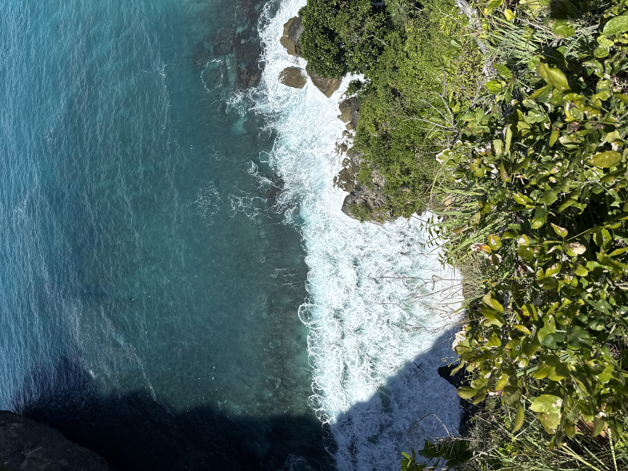 The image shows an aerial view of a coastline in Pejukutan, Indonesia. The turquoise ocean water occupies the left side of the frame, meeting the rocky shore. White waves crash against the rocks and the cliffside, creating a foamy surf. Lush green vegetation covers the cliffs, extending into the right side of the image. A shadow is cast by the cliff.