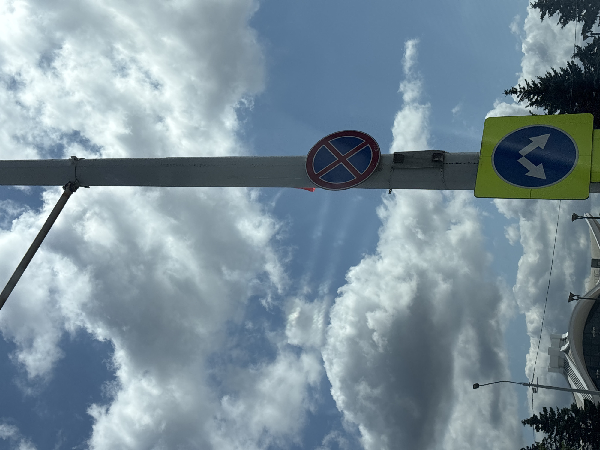 An outdoor, eye-level, slightly tilted medium shot captures a segment of a light pole against a cloudy blue sky. Attached to the pole are two circular traffic signs. The upper sign is red and blue with a white X, indicating no stopping. Below it, a yellow square sign with a blue circle and two white arrows pointing diagonally down and left suggests a mandatory turn or lane change. In the background, a modern building with a curved roof and a streetlight are visible, along with the tops of evergreen trees. The scene is set in Minsk, Belarus.