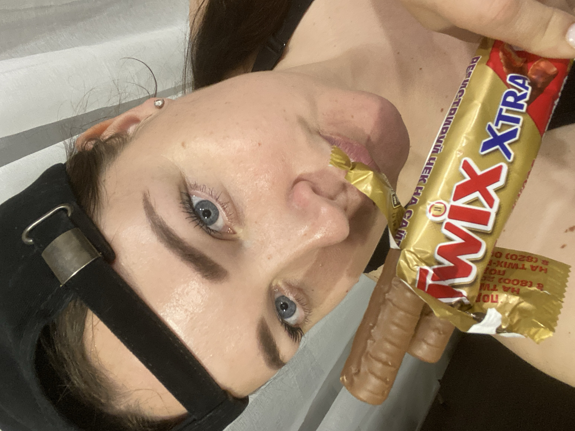 A young woman with fair skin and blue eyes is holding a Twix XTRA candy bar in her right hand and eating the candy. She is wearing a black baseball cap. The wrapper of the candy bar contains Russian text: "ВЕГИСТРИРУЙ ЧЕК НА САЙ" and the candy bar information HA TWIX-B HA (800) 20 HA TWIX-F (820). The scene is dimly lit with a gray and white background. The location is not specified.