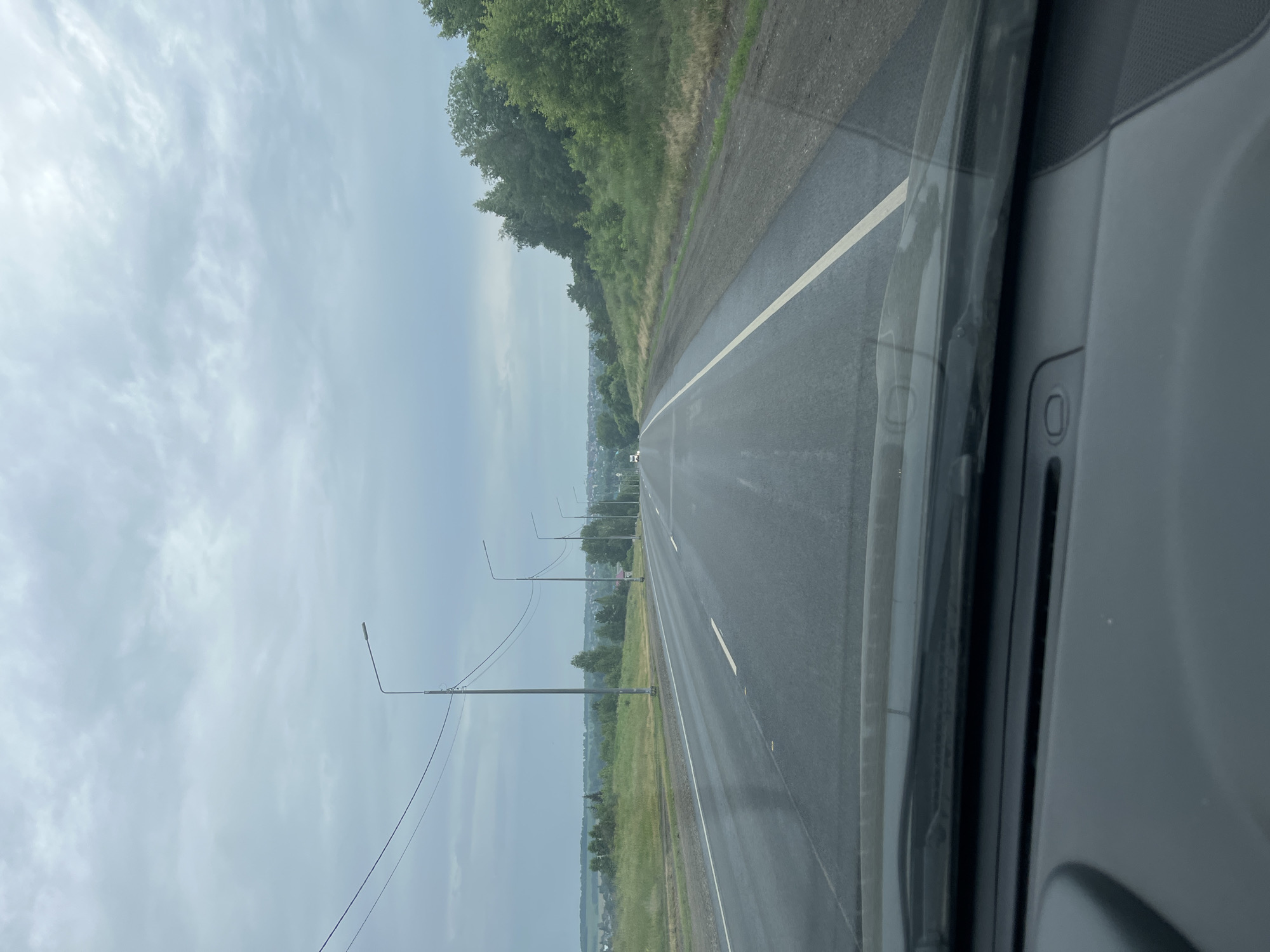 The image shows a view from inside a vehicle looking out at a two-lane asphalt road extending into the distance under a cloudy sky. The road has white lane markings and is wet, suggesting recent rain. Several streetlights are visible along the side of the road, accompanied by power lines. The landscape on either side of the road consists of grassy fields, trees, and some scattered buildings in the distance, indicating a rural or semi-rural setting. There are no visible people in the image. The front dashboard of the vehicle is partially visible in the foreground.