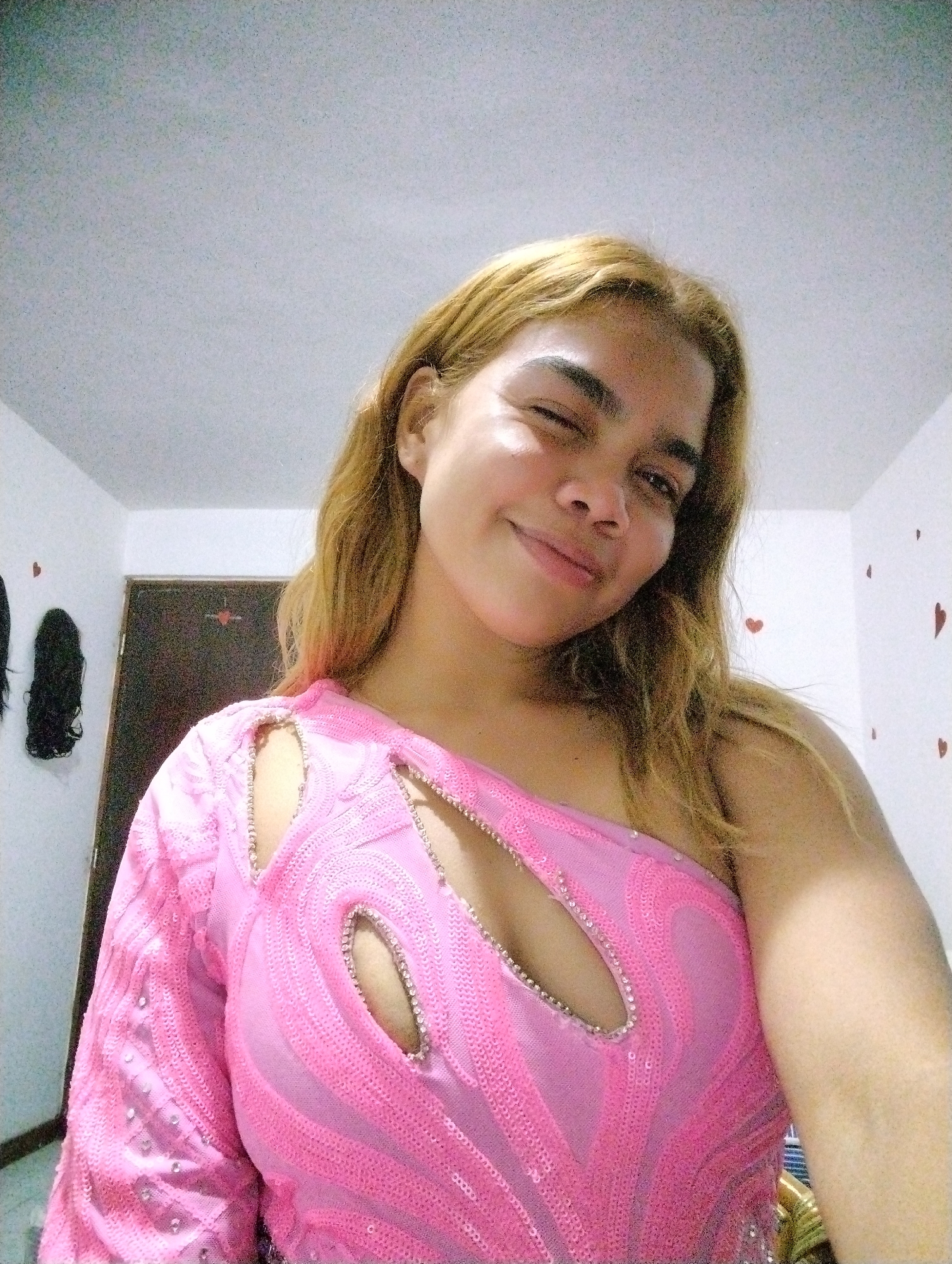 A person with light brown or blonde hair and a fair complexion is captured in a close-up, upward-angled selfie, winking with their left eye and smiling. They are wearing a bright pink, one-shoulder top embellished with sequins arranged in wavy patterns. The top features several stylized cutouts across the chest and shoulder, outlined with small, shiny beads or rhinestones. The left sleeve of the top is long and also covered in sequins.
The setting appears to be an indoor room with plain white walls. On the left side, two dark, long, wig-like objects are hanging against the wall. A dark brown or black door is partially visible in the background, with a small star-shaped decoration above it. Several red heart-shaped stickers are sparsely placed on the white wall to the right. The lighting is soft and consistent, suggesting artificial indoor illumination. The person's expression conveys a playful and cheerful mood. No text is visible in the image.
