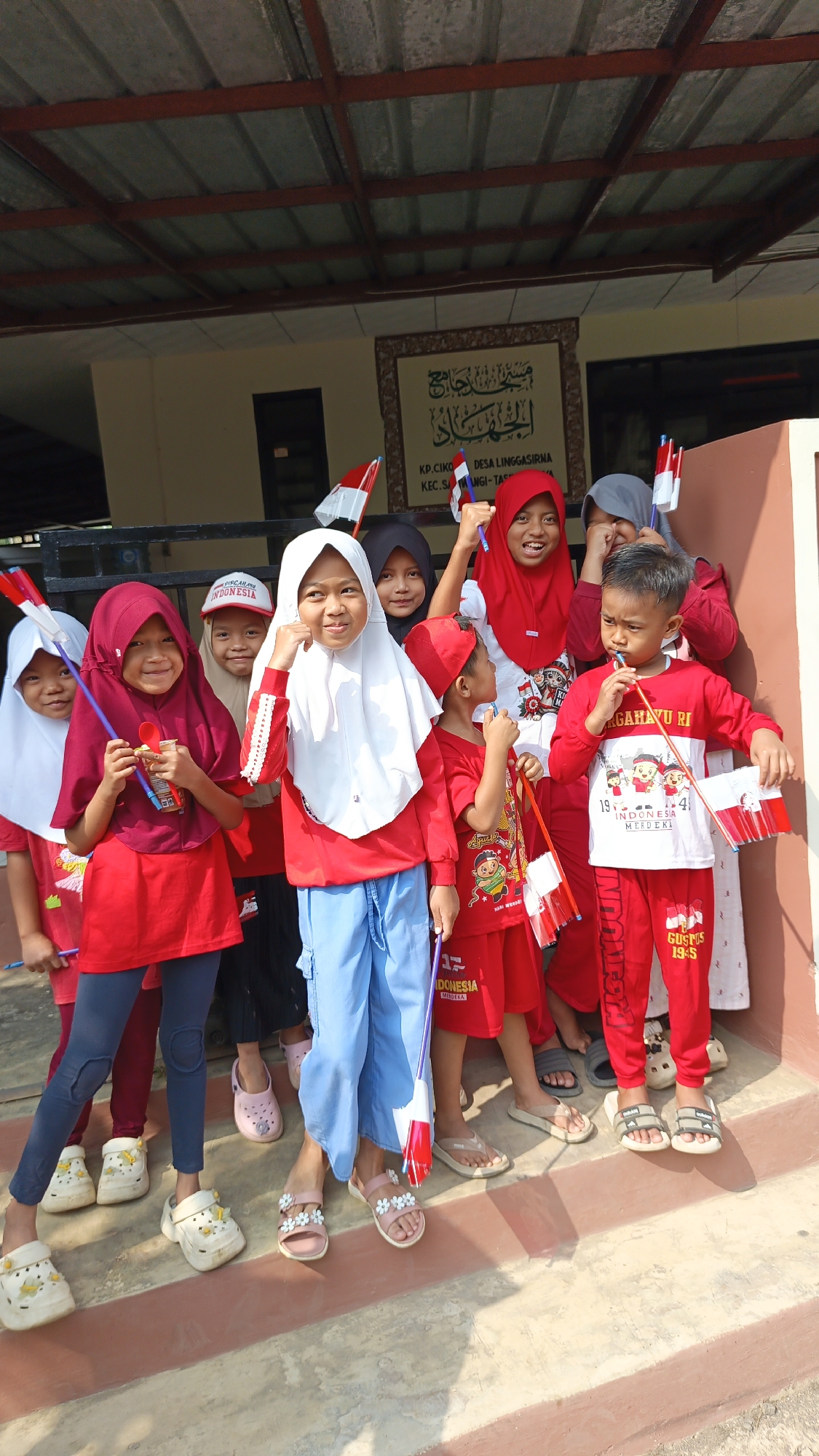 A group of eight Indonesian children, all wearing red and white attire, are posing for a photograph outdoors. The children are holding small Indonesian flags and appear to be celebrating. The background includes a building with a corrugated metal roof and a sign that reads "KP. CIKO DESA LINGGASIRNA KEC. SANGI-TAS". This indicates the location as Ciko Village, Linggasirna, in the Sangi-Tas district of Indonesia. The children are standing on steps leading up to the building. The overall atmosphere is festive and patriotic.