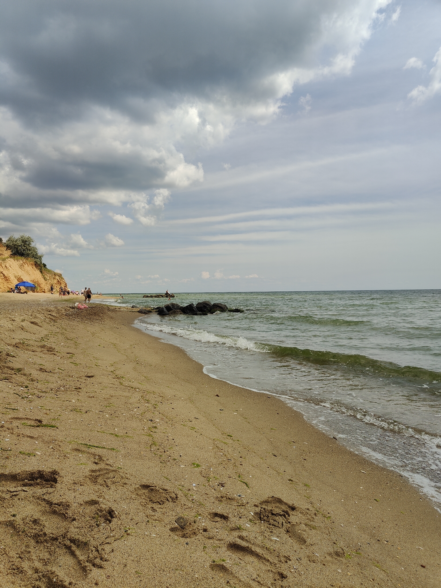 The image depicts a beach scene in Sanzhiika, Ukraine, under a partly cloudy sky. The foreground is dominated by a sandy beach with footprints and scattered small shells and seaweed. The sea is to the right, with gentle waves lapping the shore. In the middle ground, there is a breakwater made of tires filled with rocks, and further out, a person is visible in the water. The background shows a sandy cliff on the left, with some vegetation on top, and several people walking or relaxing on the beach. There is also a blue umbrella set up on the sand. The overall atmosphere is that of a typical summer day at the seaside.