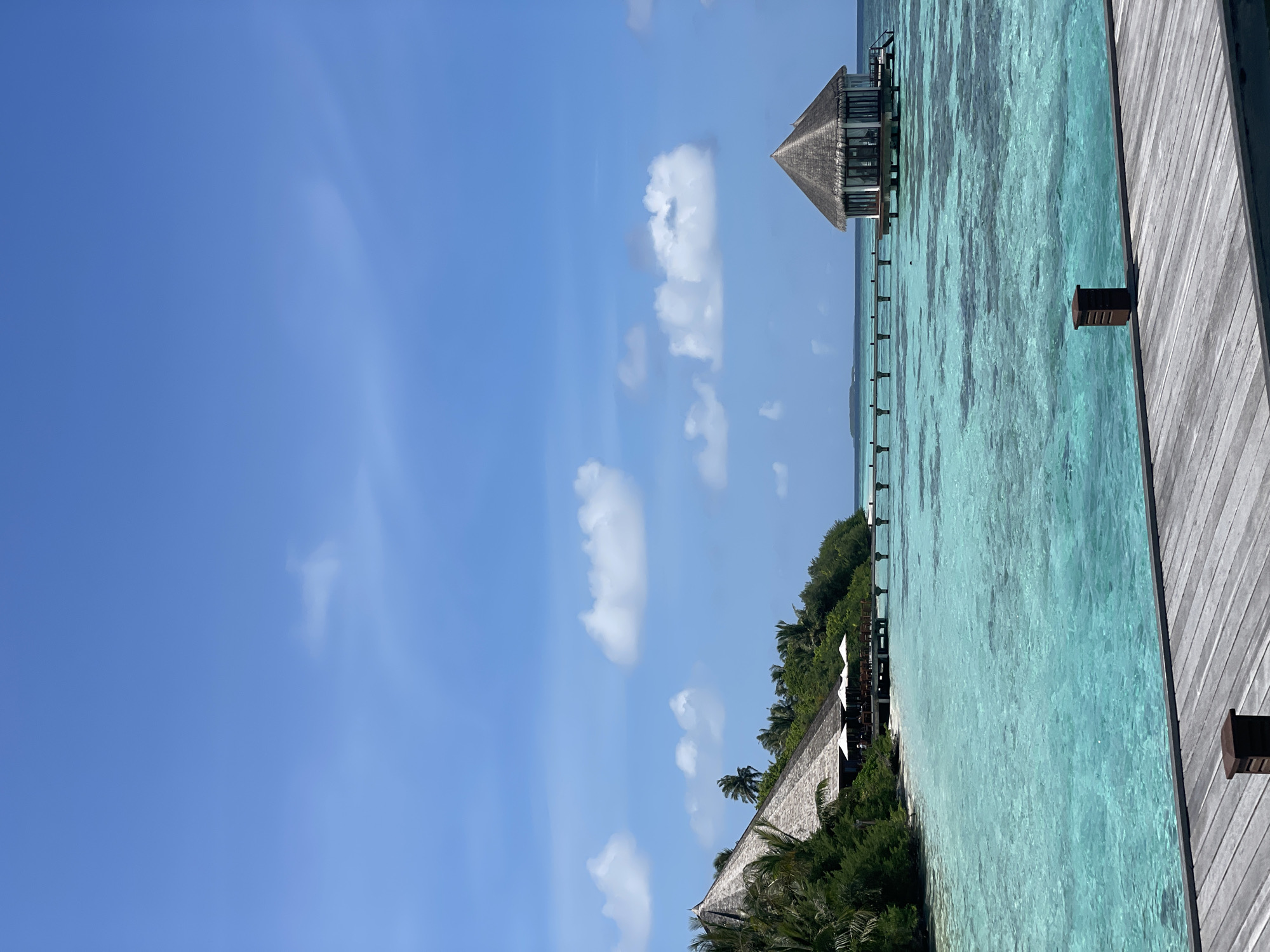 A bright, sunny day in a tropical location shows a wooden pier in the foreground. The pier is made of weathered gray planks and has a small brown wooden post on it. Beyond the pier, a body of turquoise water stretches out, with visible coral reefs beneath the surface. To the left, a lush green island rises from the water, featuring palm trees and dense foliage. On the island, a building with a thatched roof and outdoor seating with umbrellas is visible. A wooden walkway or pier extends from the island over the water towards a small, octagonal structure with a thatched roof, also situated on stilts in the water. The sky above is a clear blue with scattered white clouds. The overall scene depicts a serene and idyllic tropical resort.