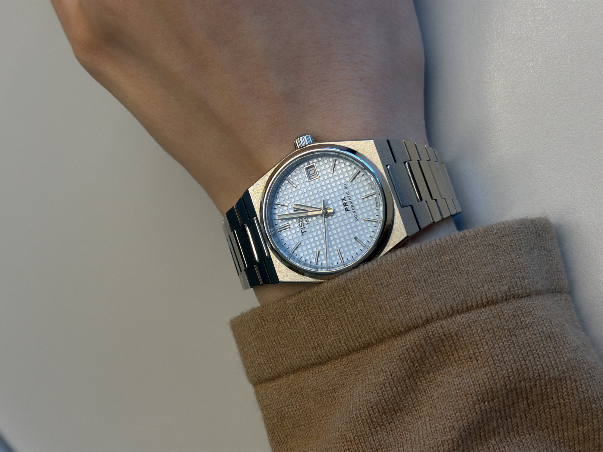 The image shows a close-up of a silver Tissot PRX Powermatic 80 watch on a person's wrist. The watch face is light blue with a textured pattern and a date window. The watchband is stainless steel and integrated into the case. The person is wearing a brown knitted sleeve. The background consists of a pale, neutral surface. There is no information to determine the location as Riga, Latvia.