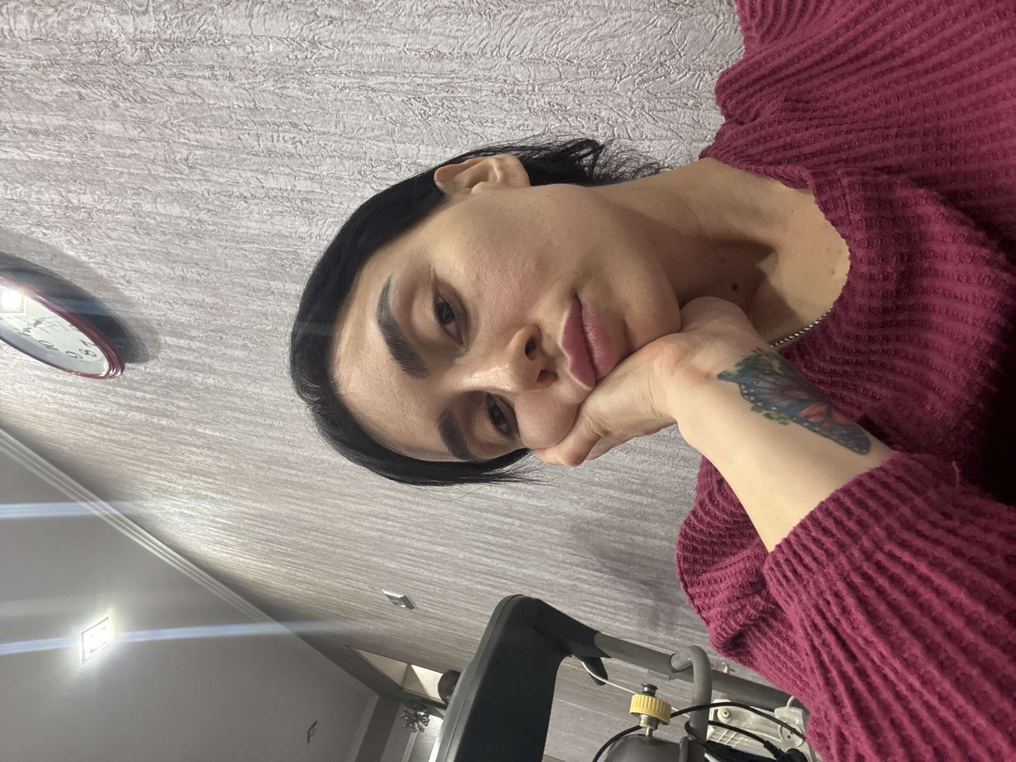 The image portrays an adult female subject indoors, located in what appears to be a residential setting within Tbilisi, Georgia. The subject has dark hair, which is pulled back from her face, and wears a magenta-colored, textured long-sleeved top. She is captured from a slightly low angle, resting her chin on her right hand, with a neutral to slightly pensive expression and a subtle pout. A colorful butterfly tattoo is visible on her left forearm/wrist, and a thin silver-colored chain necklace is present around her neck.
The background consists of a textured, light gray wallpaper. An oval-shaped, red-rimmed analog wall clock is mounted on the wall in the upper right quadrant. In the upper left quadrant, an illuminated square recessed ceiling light fixture is visible, beneath white crown molding that runs along the ceiling-wall joint. To the left of the subject, a portion of dark gray fitness equipment, consistent with a treadmill, is positioned, featuring a yellow adjustment knob and white cabling. Further to the far left, an opening reveals a glimpse into an adjacent room or hallway, where a decorative light fixture and a lighter-colored wall are discernible.