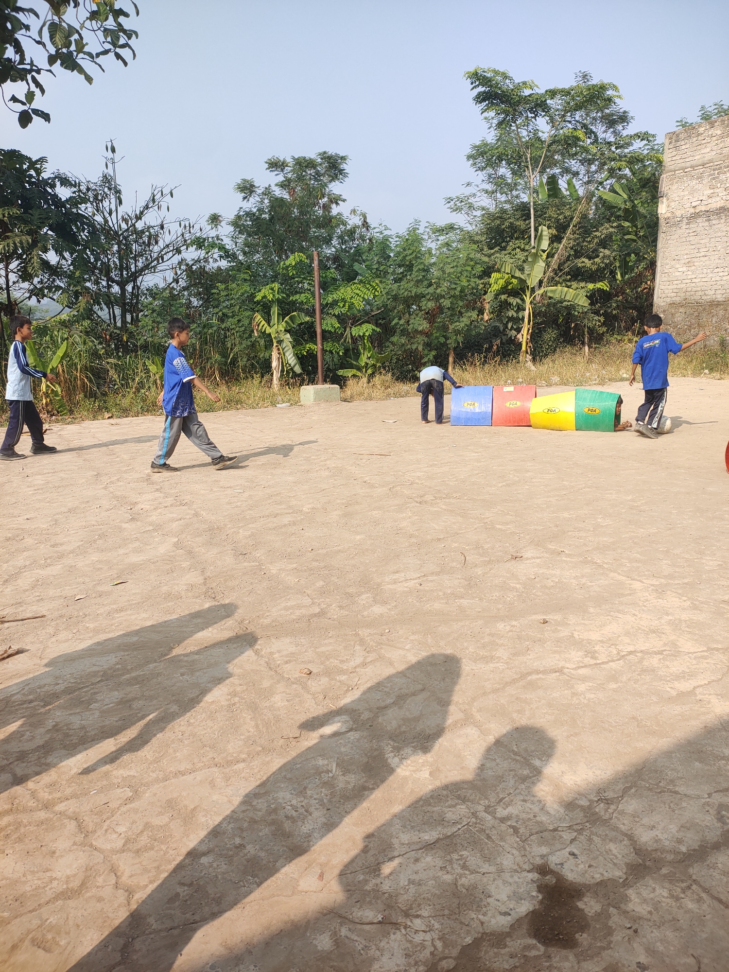 In an outdoor, sunlit, dirt-grounded area, four boys are engaged in what appears to be a game. Two boys are in the foreground, walking left and right, their shadows cast prominently on the ground. In the mid-ground, a boy in a blue shirt is kicking a white soccer ball. To his left, another boy in a blue shirt is partially obscured, possibly interacting with colorful, modular boxes that are lined up. The background features lush green trees, foliage, and a brick structure, suggesting a rural or semi-rural setting within Subang, Indonesia. The sky is clear and light blue.