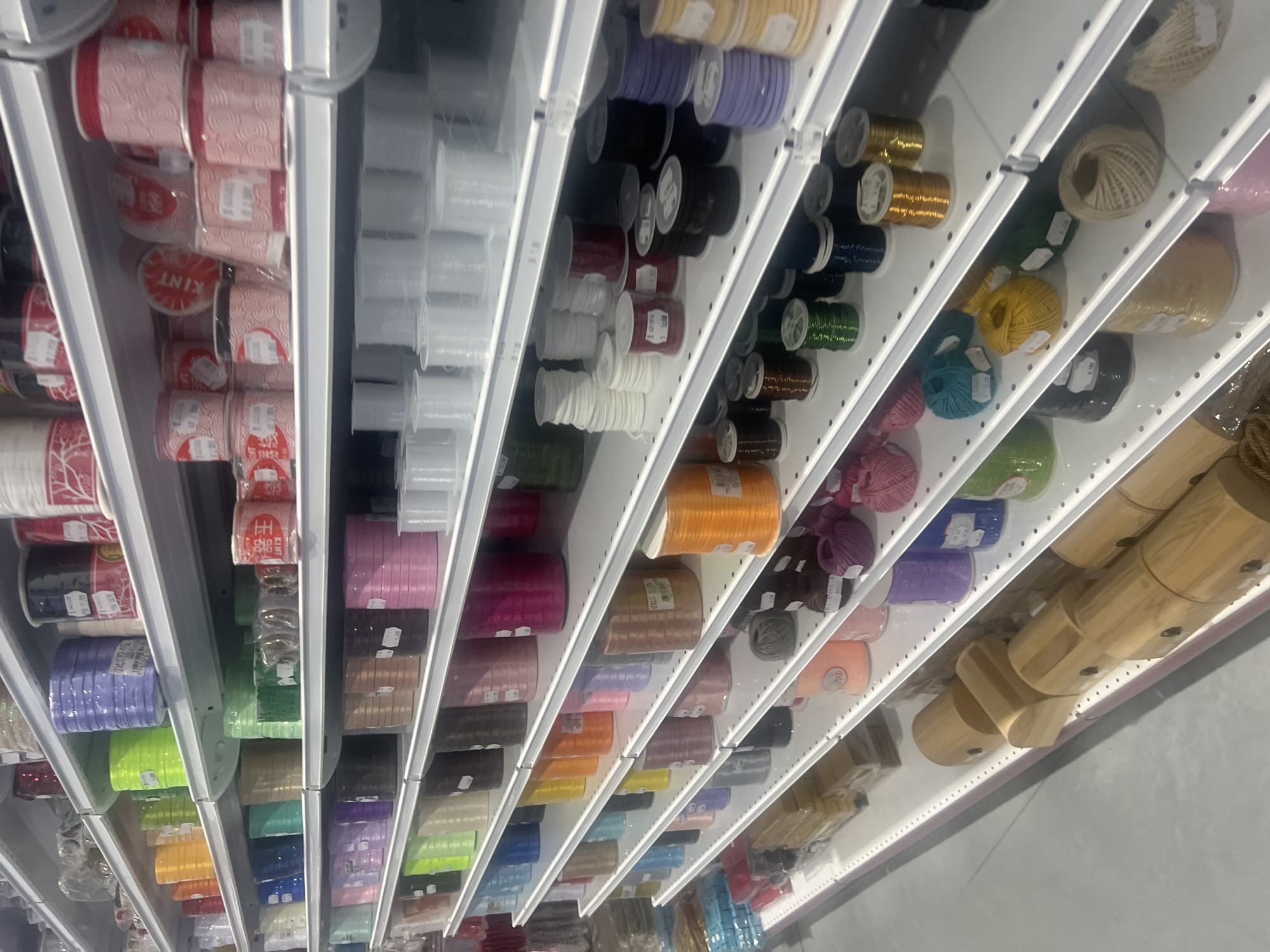 The image shows shelves filled with spools of ribbon and yarn of various colors and textures in a retail setting. The scene appears to be inside a craft or fabric store, possibly in Burgas, Bulgaria, given the request. The spools are neatly arranged on white shelves. From left to right, the colors and materials range from red ribbons, transparent plastic spools, and a spectrum of colored yarns. Wooden objects are located on the rightmost shelf.