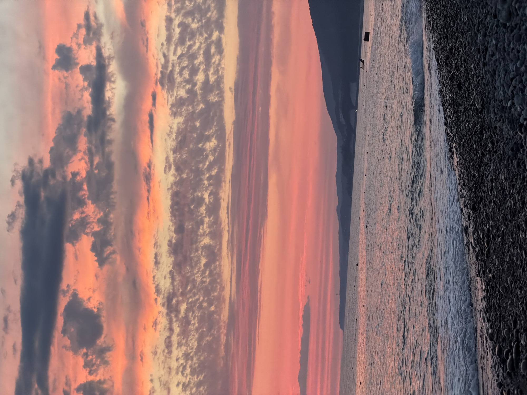 The image depicts a serene sunset over the Black Sea coastline in Sochi, Russia. The sky is a vibrant canvas of pinks, oranges, and purples, painted with layers of clouds. In the foreground, a pebbled beach transitions into the gentle waves of the sea. The distant horizon is marked by a dark, silhouetted coastline with rolling hills. The water reflects the warm hues of the sunset, creating a tranquil atmosphere. There are no people visible in the image. A few small objects, possibly buoys or markers, are present in the water. The overall scene captures the natural beauty of a coastal evening.