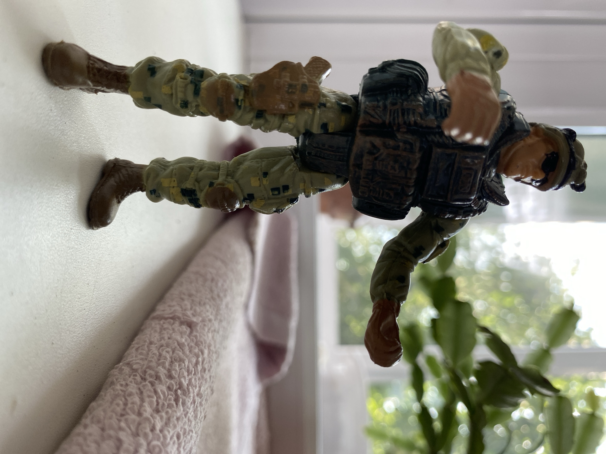 A full-body, eye-level, low-angle shot shows a plastic toy soldier standing on a white surface. The soldier is wearing camouflage fatigues, a tactical vest with pouches, sunglasses, and a helmet. The soldier's right arm is extended forward, and the left arm is bent at the elbow, with the hand open as if reaching for something. A holstered pistol is attached to the soldier's right thigh.
In the background, a blurred potted plant with green leaves is visible in front of a window, suggesting an indoor setting. A rolled-up pink towel lies on the white surface to the right of the soldier. The scene appears to be a still life arrangement, possibly on a windowsill or table. There is no indication of the city of Бескөл ауылдық округі, Kazakhstan within the image.